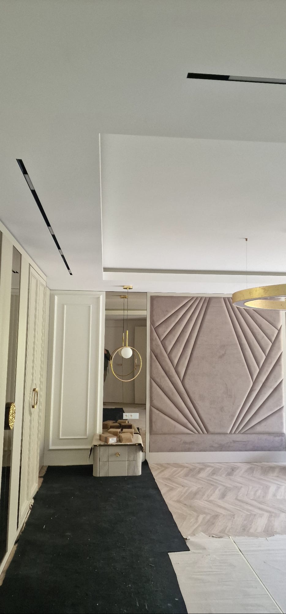 Nowoczesna sypialnia w trakcie wykończenia: tapicerowany panel ścienny z geometrycznym wzorem, parkiet w jodełkę, lustro i minimalistyczne oświetlenie.