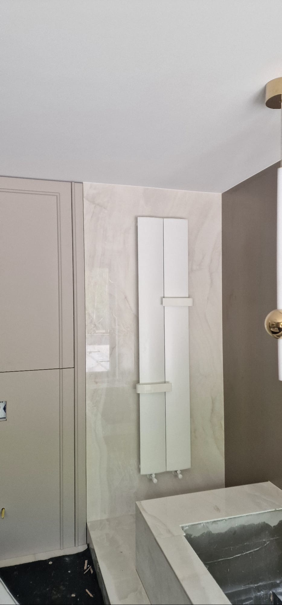Biała szafka z grzejnikiem łazienkowym na marmurowej ścianie, obok beżowej szafki i lampy. Widok na fragment wykończonej łazienki po remoncie.