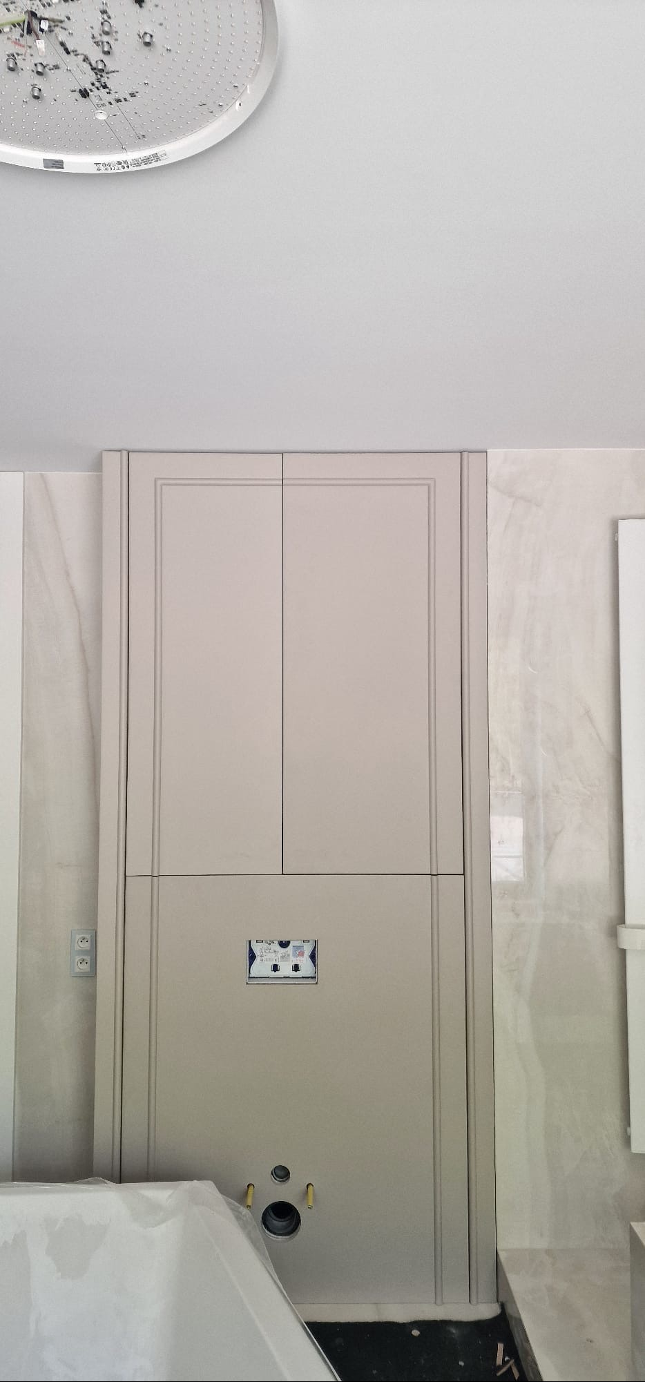 Szara zabudowa w łazience z ukrytymi drzwiczkami rewizyjnymi i elementami instalacji, marmurowe ściany, widoczny fragment wanny zakrytej folią, sufit z oświetleniem.