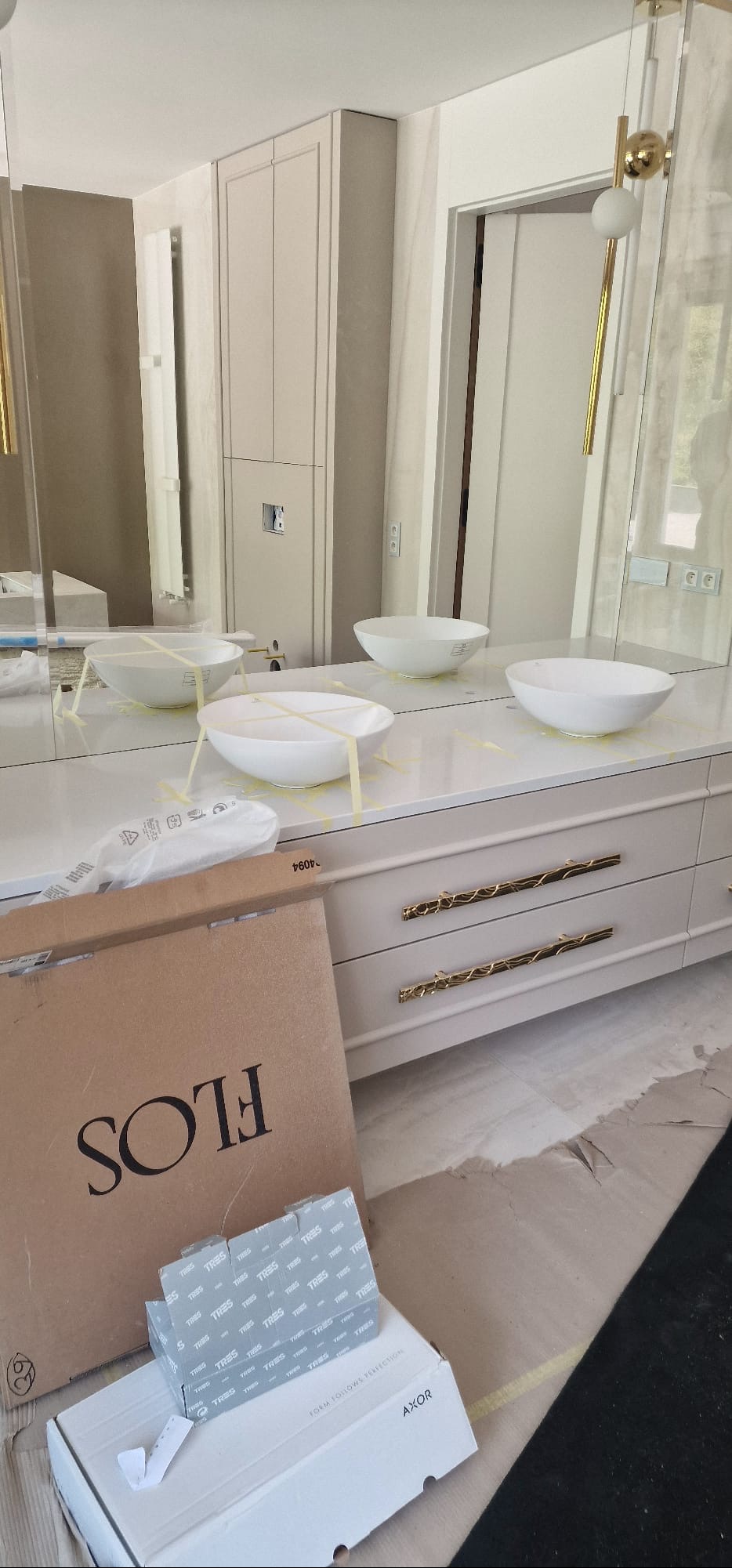 Nowoczesna łazienka w trakcie montażu: biała szafka z umywalkami, złote uchwyty, kartony FLOS i AXOR. Umywalki zabezpieczone taśmą. Widoczne elementy oświetlenia.