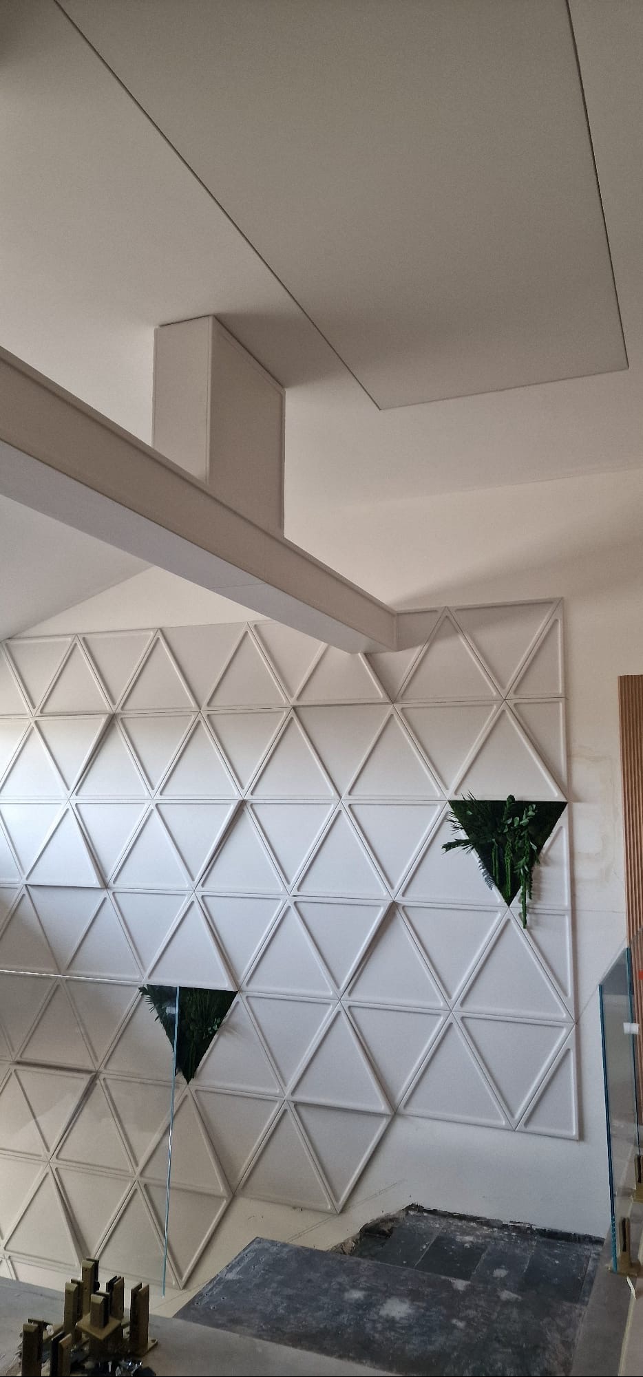 Biała ściana z trójkątnymi panelami 3D, w których umieszczono rośliny. Widoczny fragment sufitu i podłogi w trakcie prac wykończeniowych.