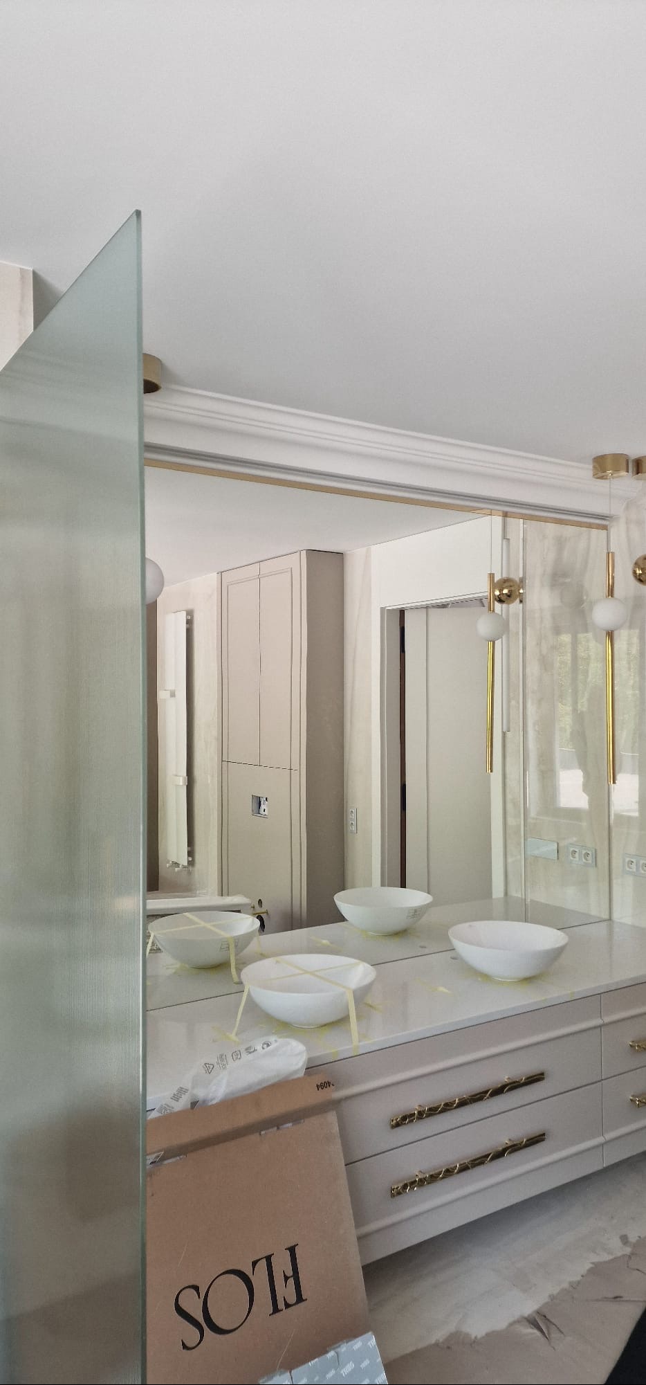 Elegancka łazienka w trakcie wykończenia: lustrzana ściana, umywalki zabezpieczone taśmą, szafki z dekoracyjnymi uchwytami, karton po lampach Flos.