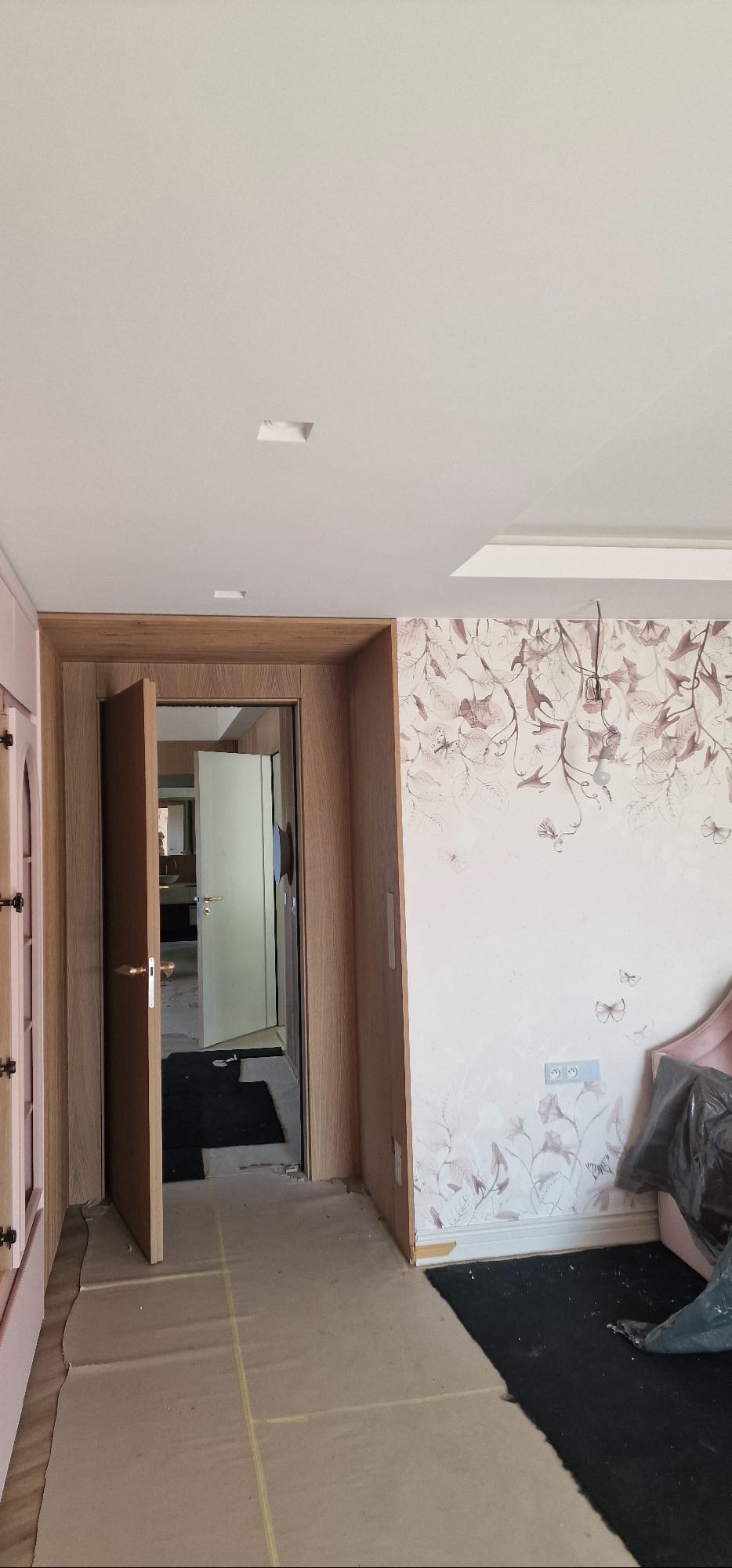 Pokój w trakcie wykończenia: ściana z tapetą w kwiaty, drewniana rama drzwi z otwartymi drzwiami i zabezpieczona podłoga. Widoczne niedokończone detale.