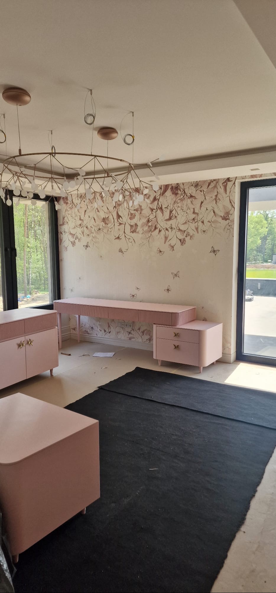 Jasne wnętrze z meblami w odcieniach pudrowego różu i tapetą w kwiaty. Widoczne duże okna, nowoczesna lampa i czarny dywan zabezpieczający podłogę.