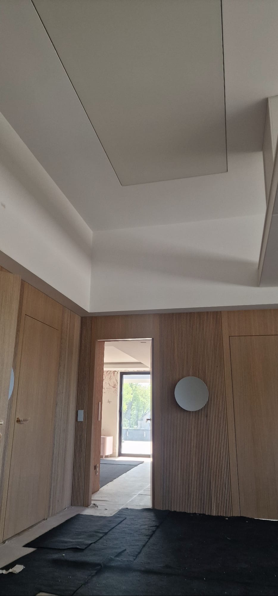 Wykończone wnętrze z drewnianą boazerią, ukrytymi drzwiami i okrągłym lustrem. Widok na jasne pomieszczenie przez otwarte drzwi. Detal wykończenia sufitu.