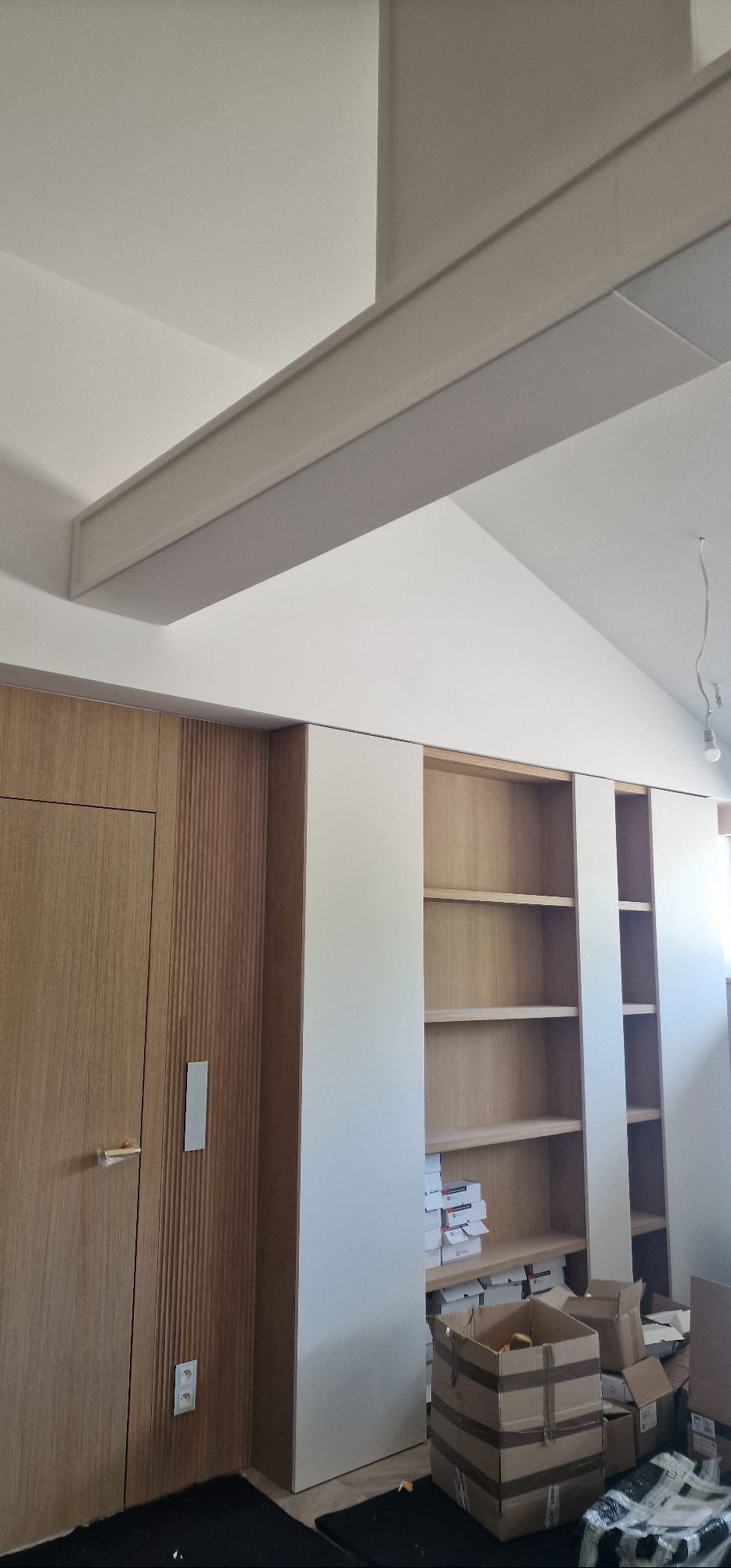 Nowoczesna zabudowa meblowa z drewnianymi elementami i białymi panelami, widoczne półki i drzwi, sufit ze skośnym elementem. Wnętrze w trakcie wykańczania.