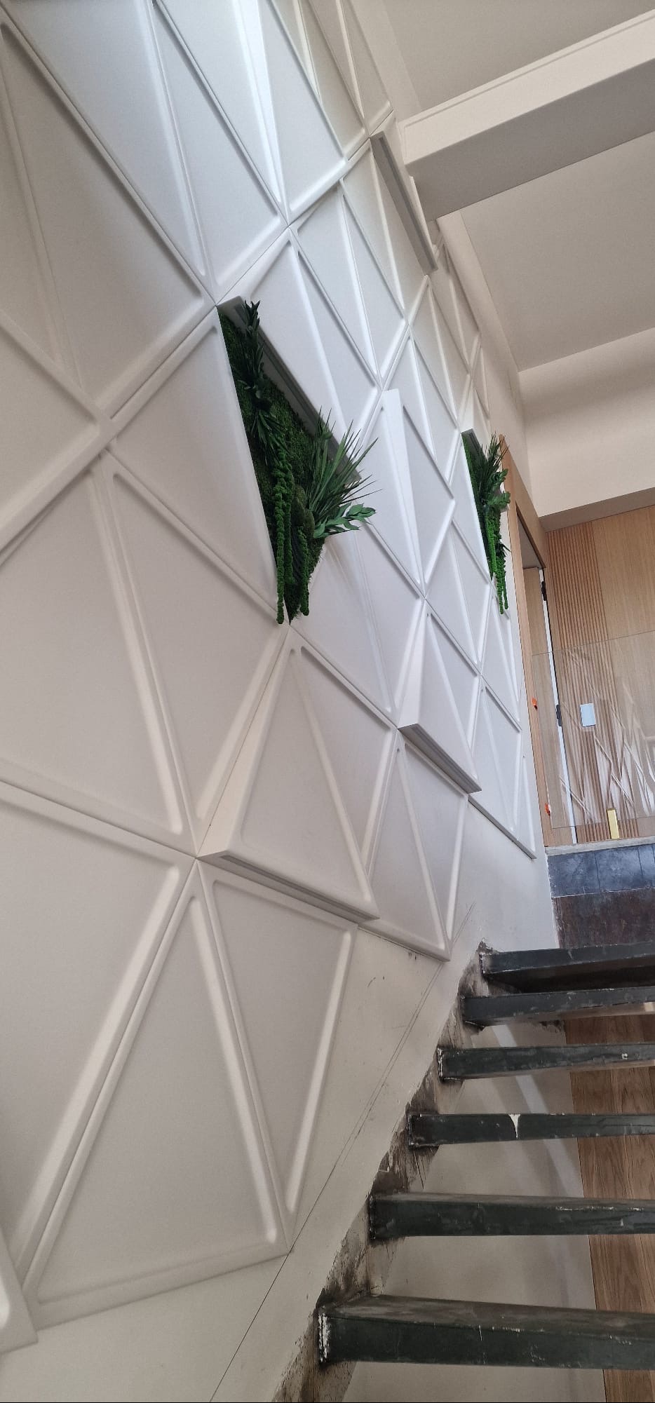 Biała ściana z geometrycznymi panelami 3D i wnękami z zieloną roślinnością, widok z dołu schodów. Nowoczesne wykończenie wnętrza.