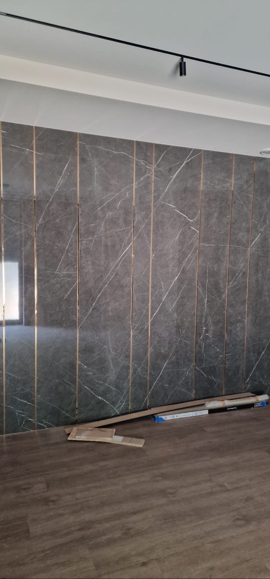 Ściana wykończona panelami z efektem marmuru, oddzielonymi złotymi listwami. Na podłodze leżą materiały budowlane, a na suficie widoczna jest czarna szyna oświetleniowa.