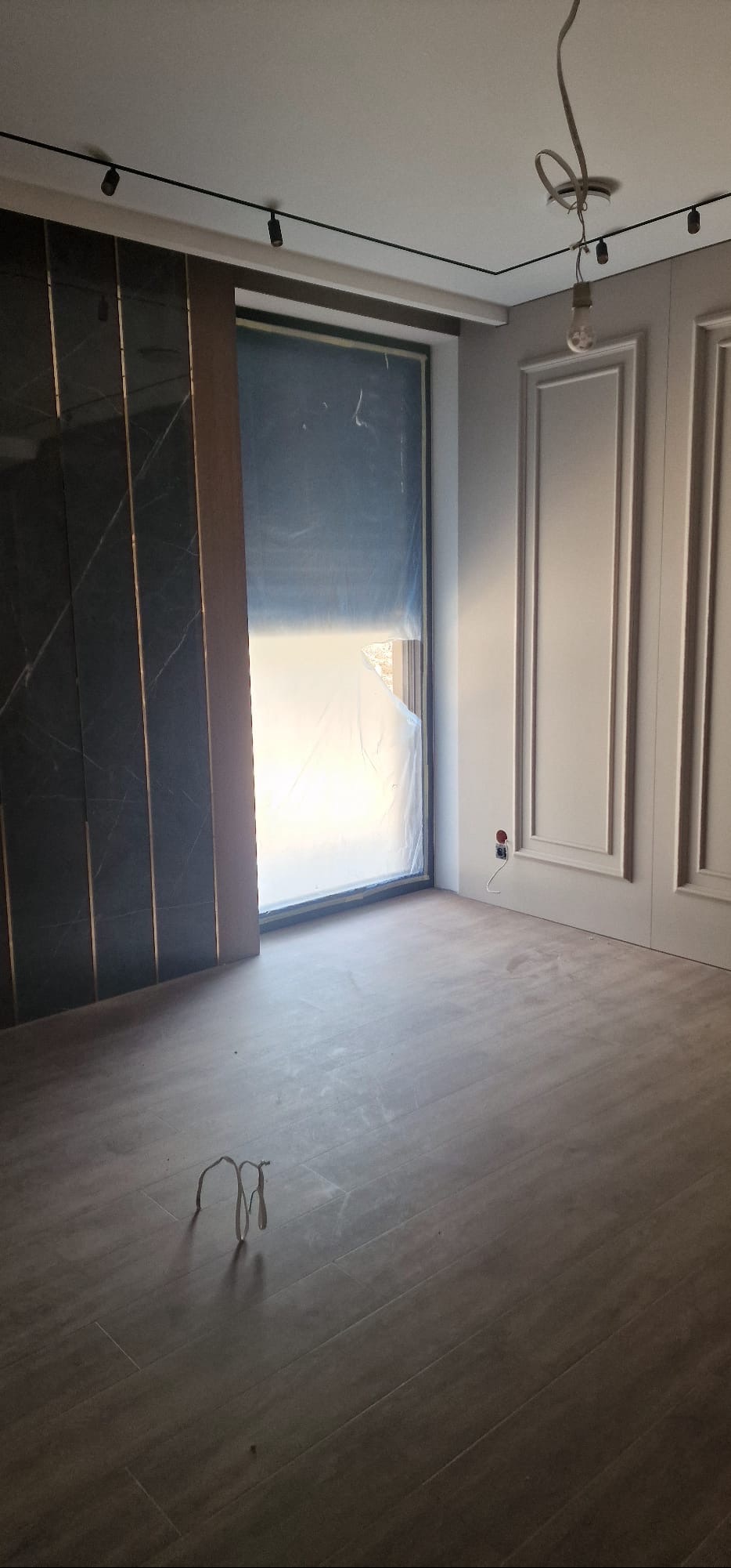 Wnętrze pokoju w trakcie remontu: położona podłoga, okno zabezpieczone folią, ściany z dekoracyjnymi panelami i listwami. Widoczne przewody elektryczne.