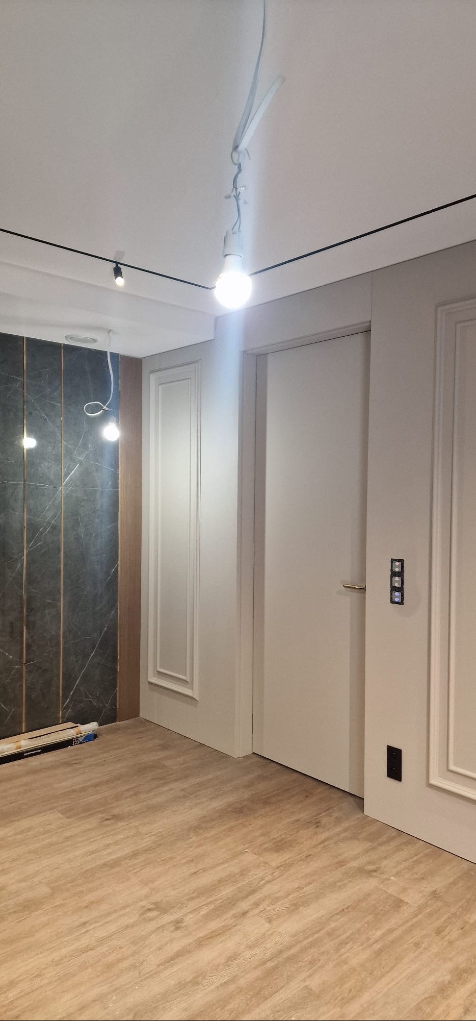 Wykończone wnętrze: drewnopodobna podłoga, dekoracyjna ściana z motywem marmuru, białe drzwi z ozdobnymi listwami i nowoczesne oświetlenie sufitowe.