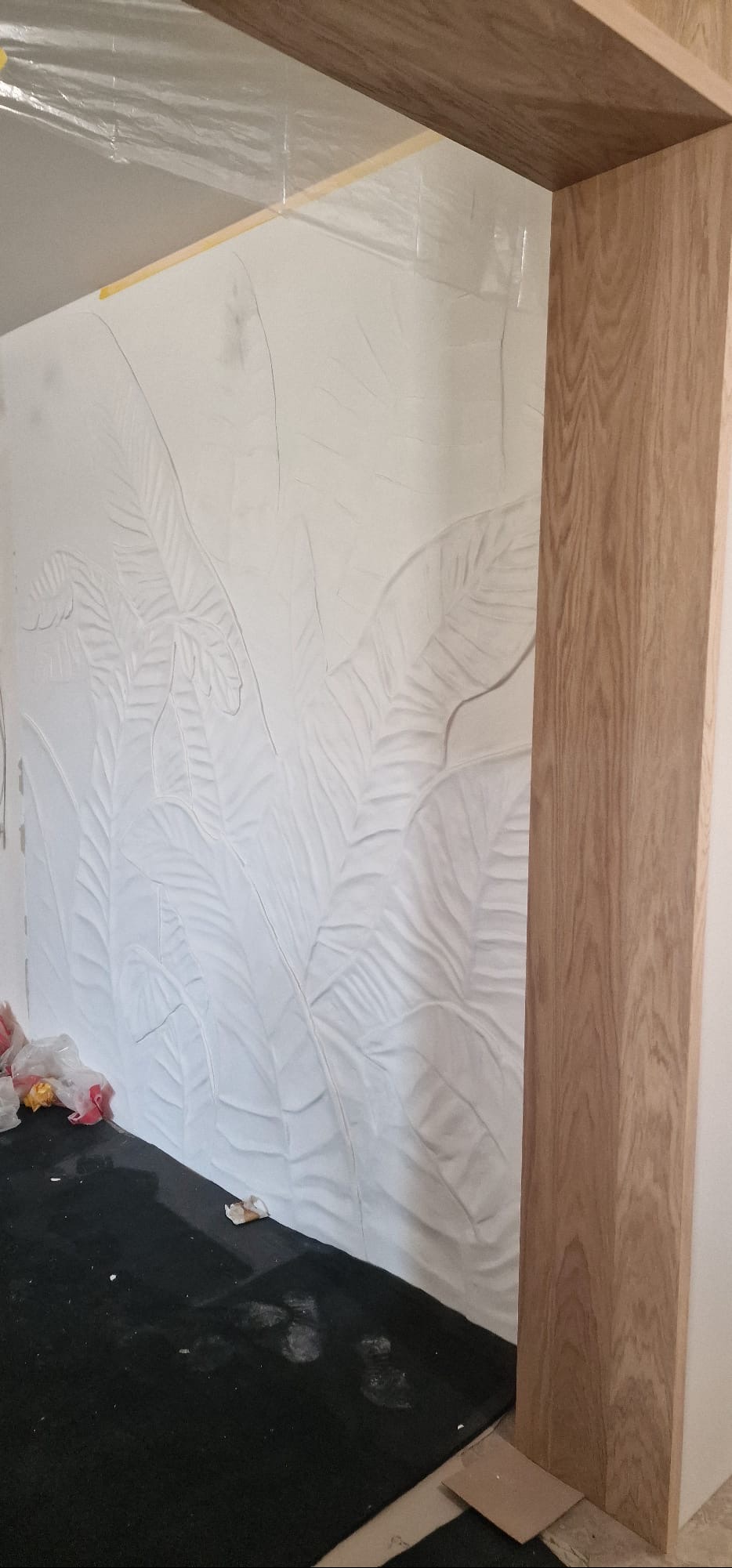 Ściana z dekoracyjnym, białym tynkiem strukturalnym w motywie liści, widoczna przez drewnianą futrynę. Podłoga zabezpieczona folią.