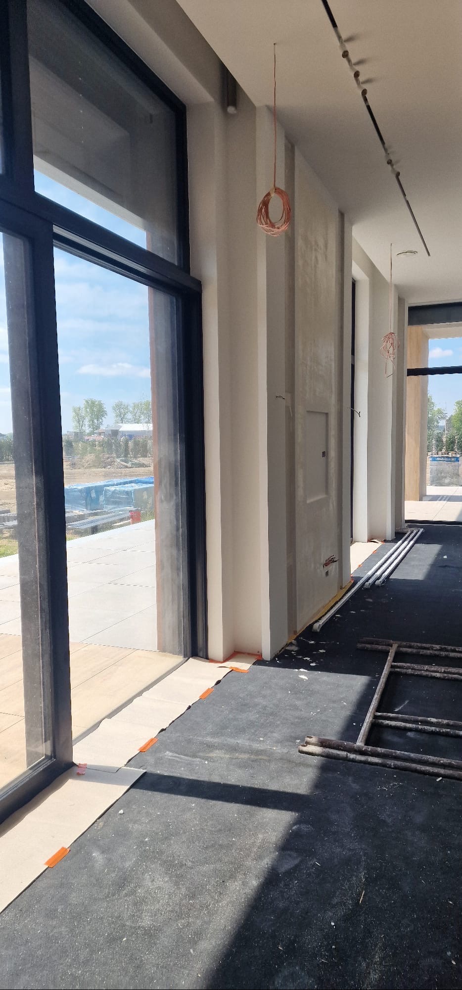 Wnętrze w trakcie wykańczania: duże okno z czarną ramą, szare ściany z widoczną fakturą, podwieszona instalacja elektryczna, zabezpieczona podłoga, drabina malarska.