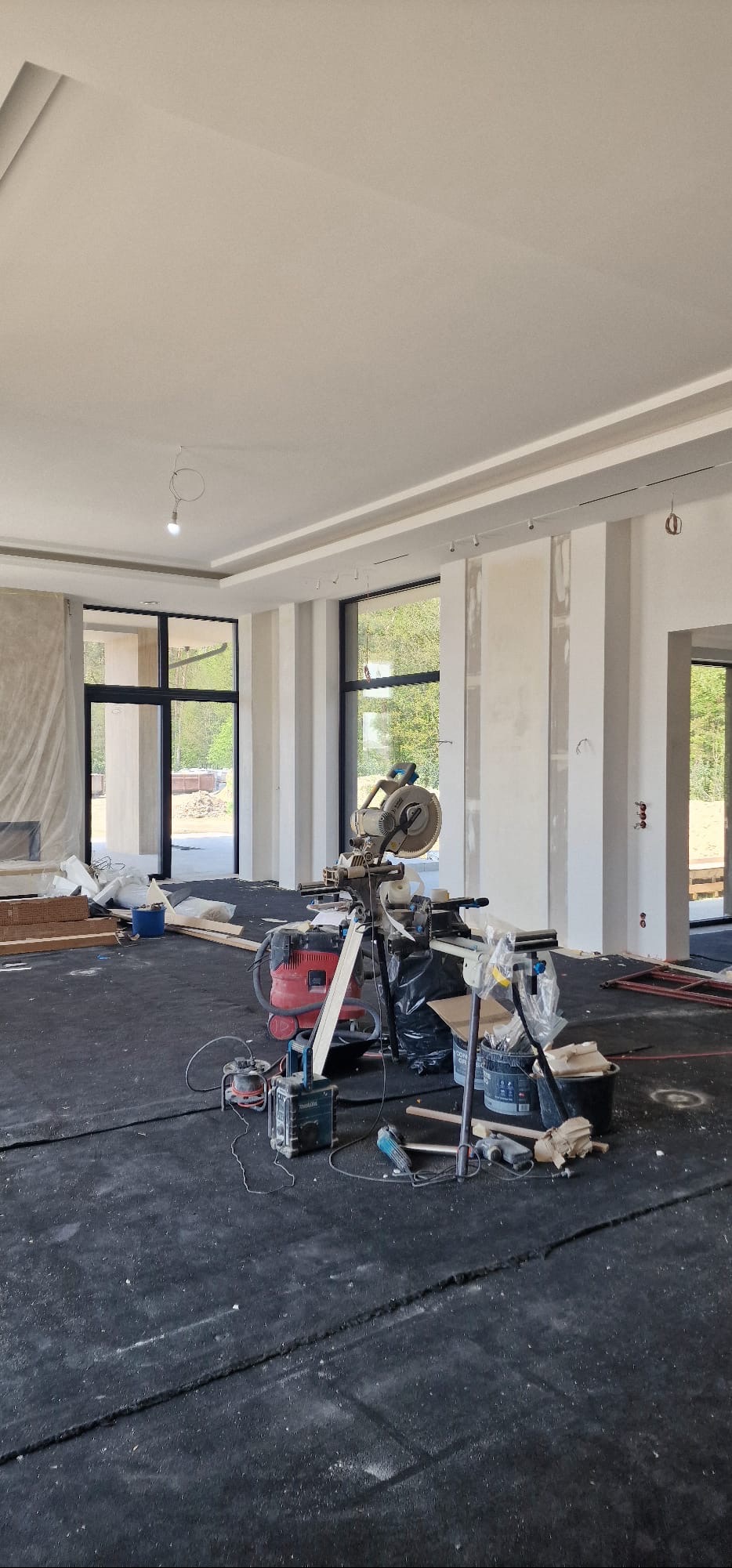 Wnętrze w trakcie wykończenia: widoczne narzędzia, w tym piła ukośnica, oraz przygotowane ściany z oknami. Na podłodze czarna folia ochronna.