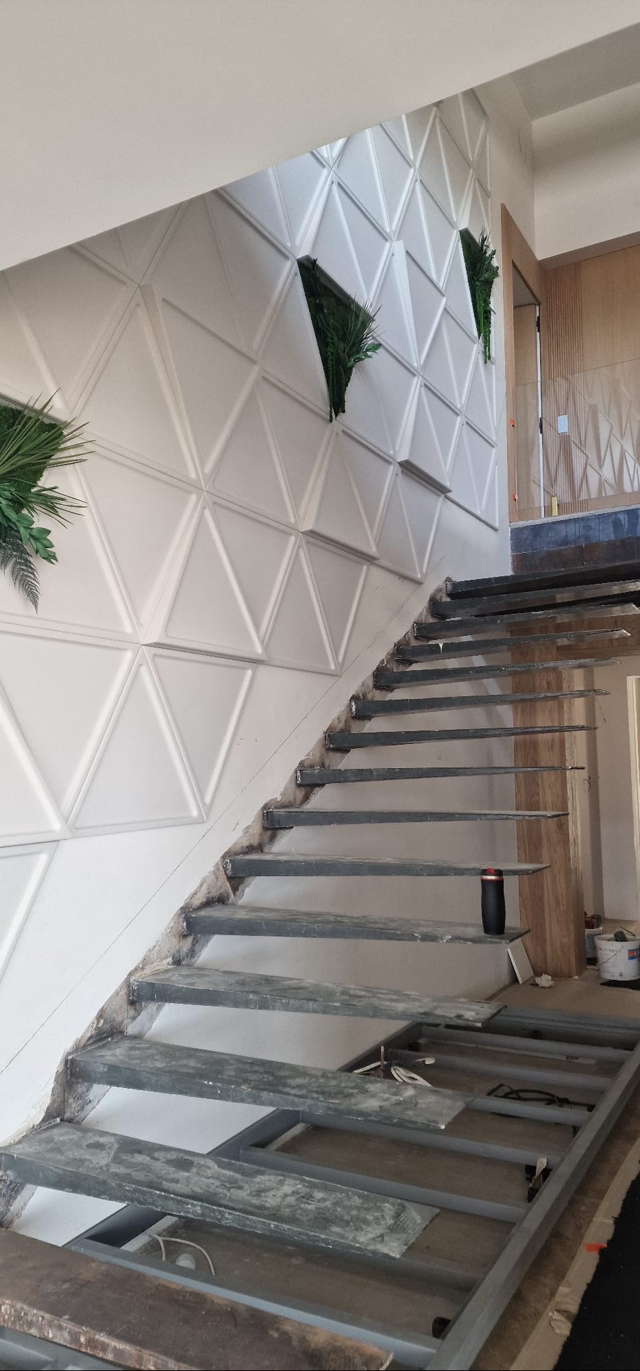 Montaż schodów z metalową konstrukcją i dekoracyjną ścianą z trójkątnymi panelami oraz zielonymi wstawkami. Wnętrze w trakcie wykończenia.