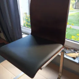 Krzesło z ciemnobrązowym oparciem i czarnym siedziskiem, na metalowych nogach. W tle okno z widokiem na ogród i narzędzia tapicerskie.