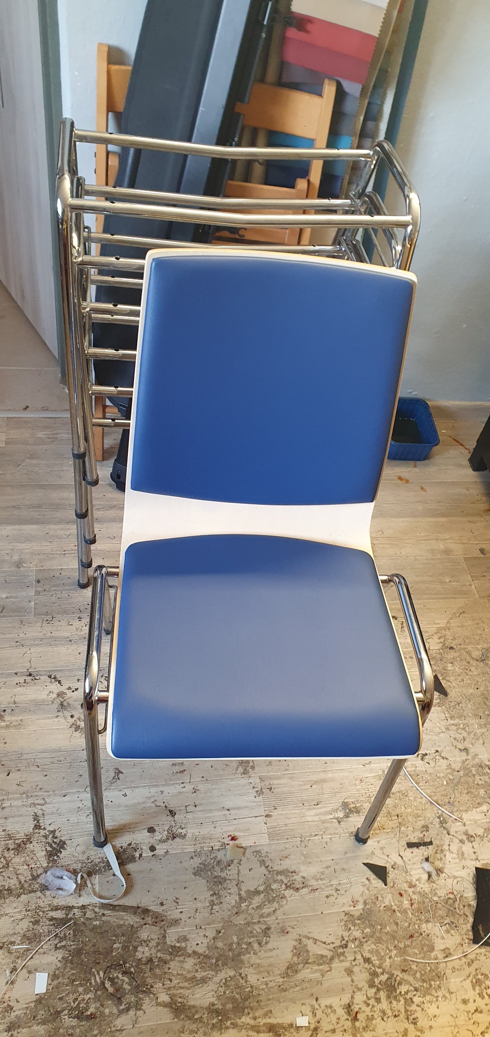 Krzesło biurowe z niebieskim siedziskiem i oparciem, na metalowym stelażu. Na podłodze widoczne ślady pracy, w tle ustawione inne krzesła.