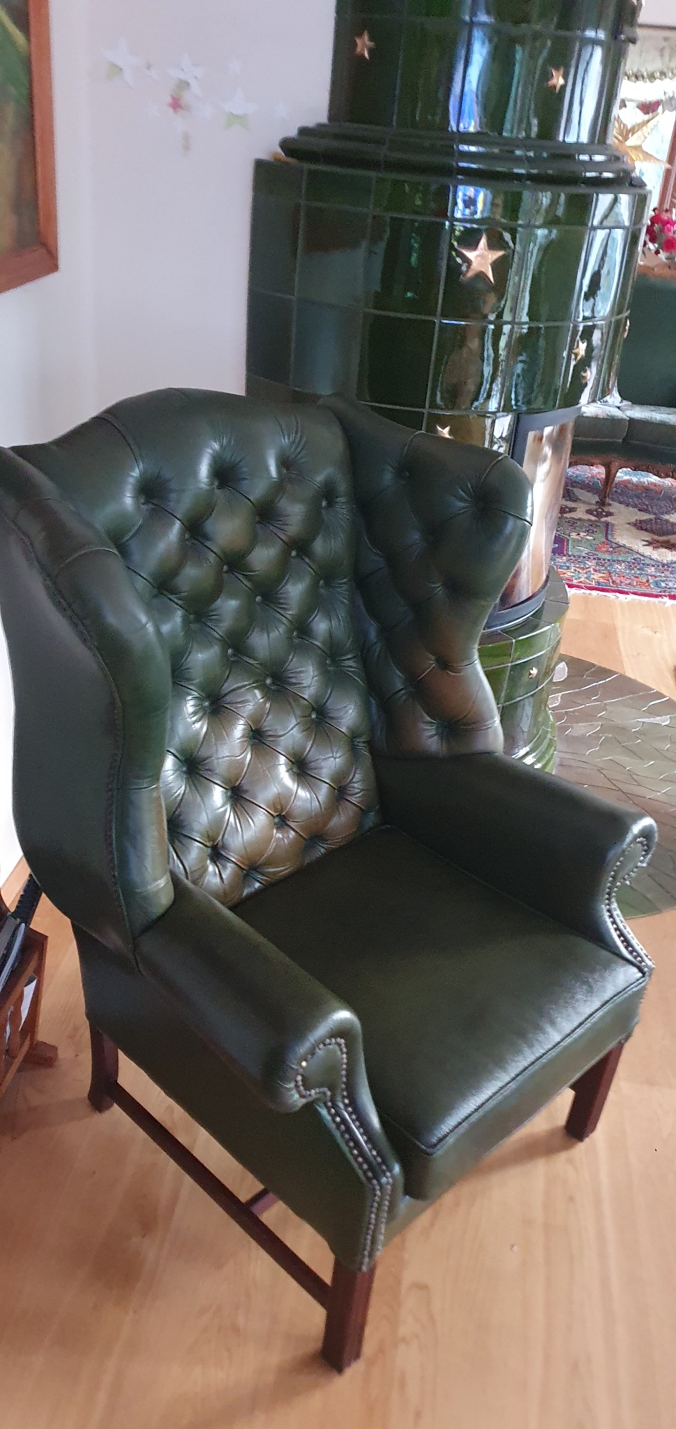 Zielony, skórzany fotel typu uszak z pikowanym oparciem, ustawiony na drewnianej podłodze, obok pieca kaflowego. Widoczne zdobienia nitami na podłokietnikach.
