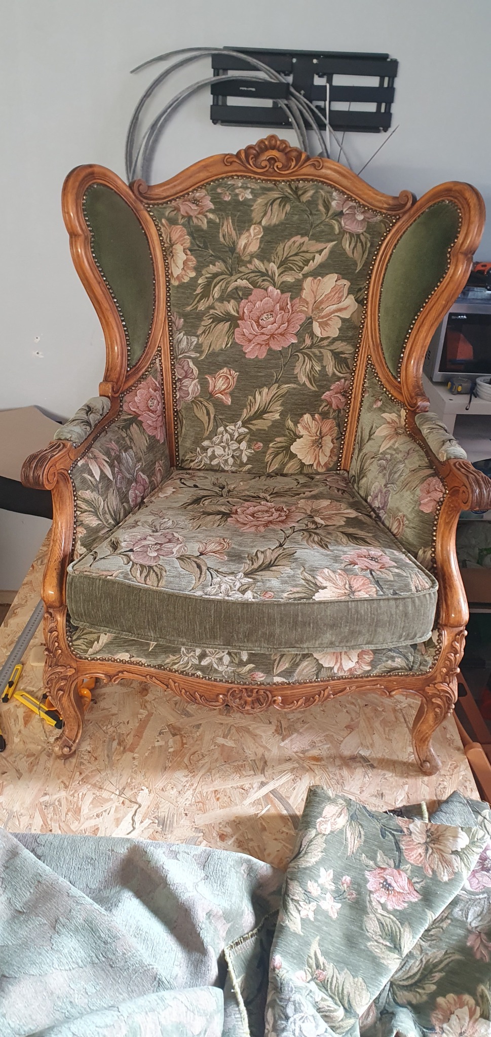 Antyczny fotel z drewnianą ramą, obity zieloną tkaniną w kwiaty, stoi na drewnianym podłożu. Widoczny fragment tkaniny i narzędzia tapicerskie.
