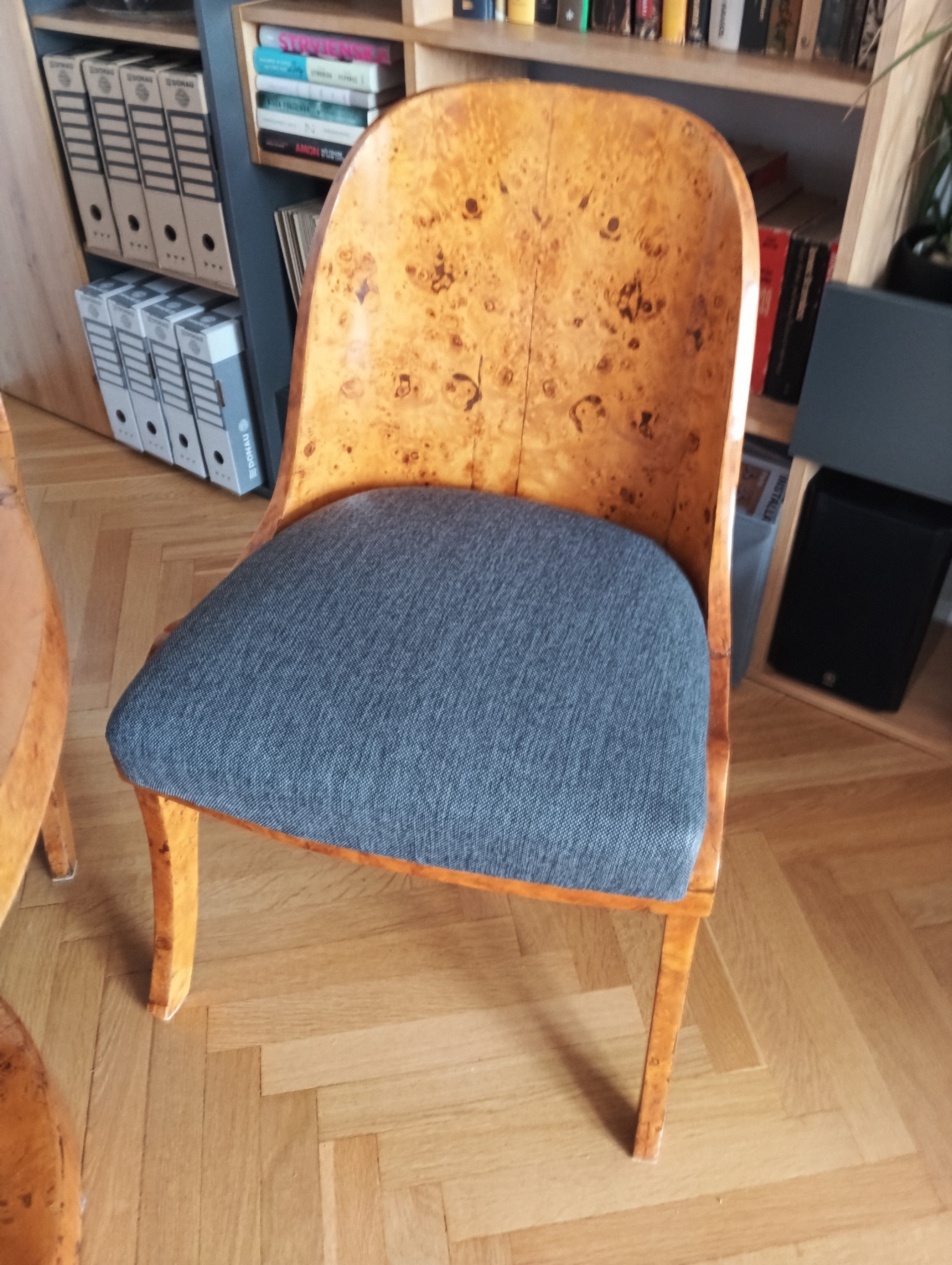 Krzesło po renowacji w Warszawie: drewniane, z tapicerowanym szarym siedziskiem. Widoczne unikalne wzory słojów drewna i odnowiona tapicerka na tle regału z książkami.