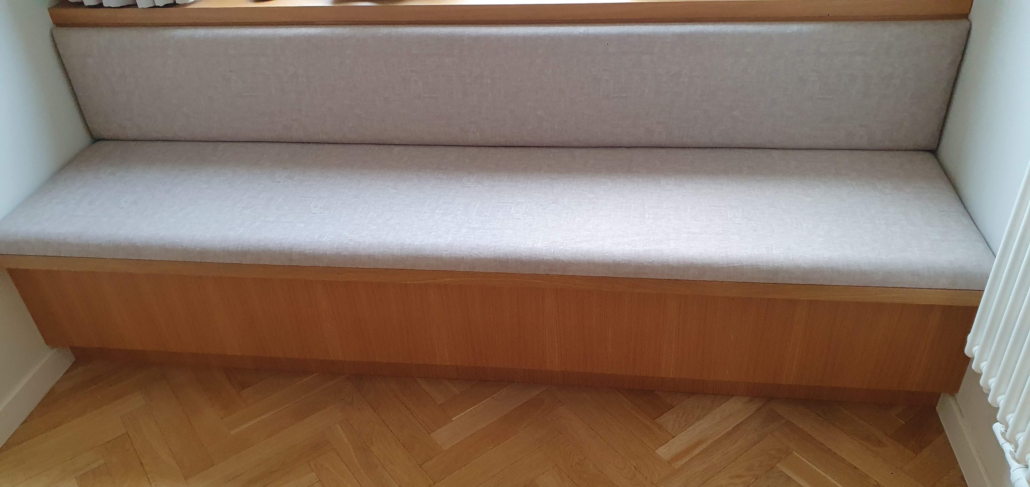 Wnęka okienna z siedziskiem i tapicerowanym oparciem w jasnym odcieniu. Parkiet w jodełkę na podłodze. Widoczny fragment grzejnika.