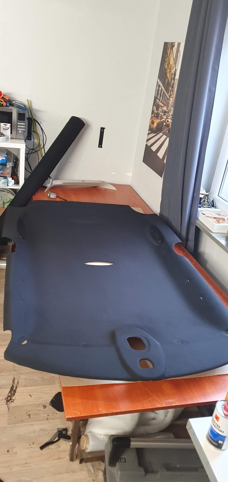 Ciemnoszara podsufitka samochodowa leży na stole w warsztacie, widoczne otwory montażowe i rolka materiału tapicerskiego w tle. Na ścianie plakat z Nowym Jorkiem.