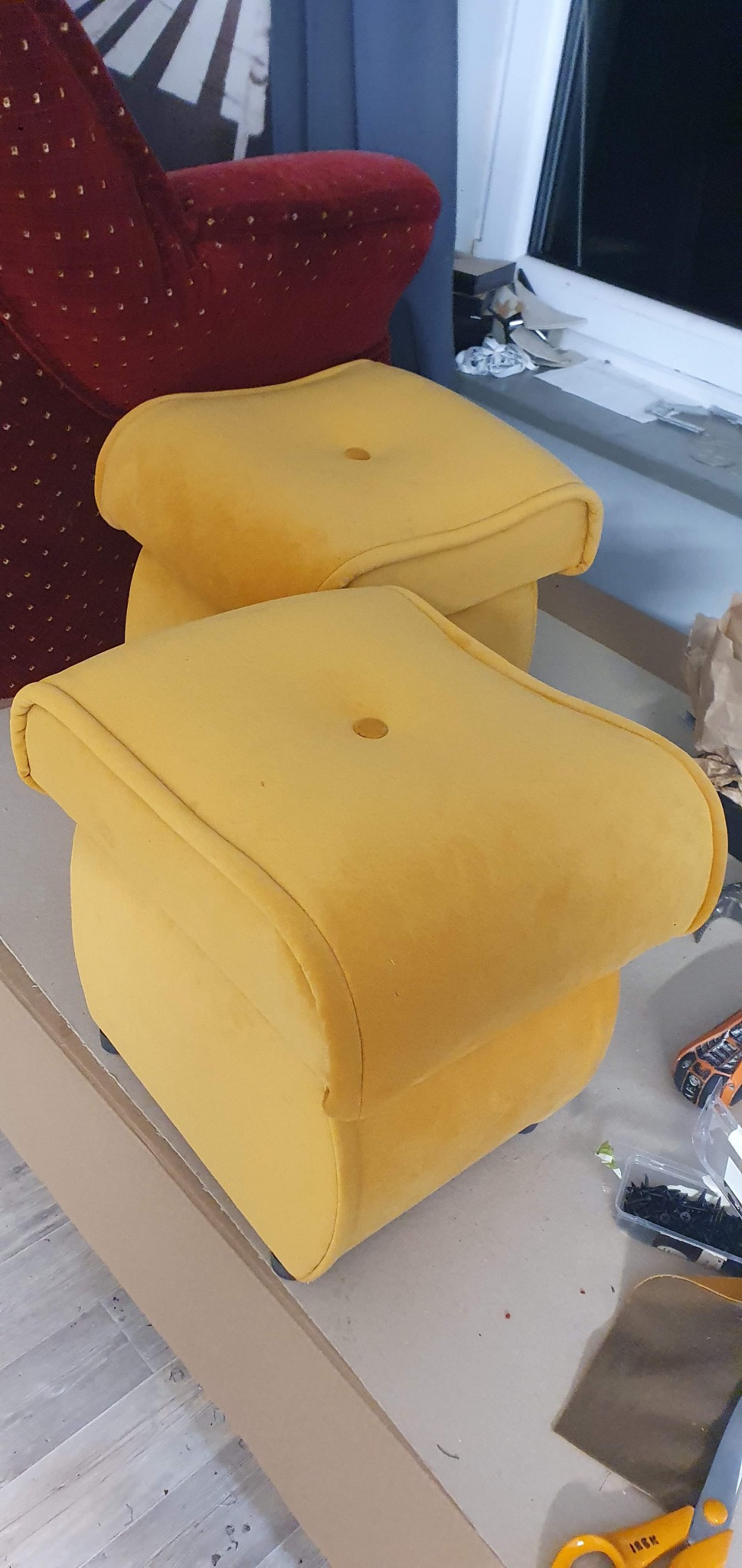 Dwa żółte, tapicerowane pufy z guzikiem, ustawione jeden za drugim. W tle widoczne krzesło i okno. Na stole narzędzia tapicerskie.