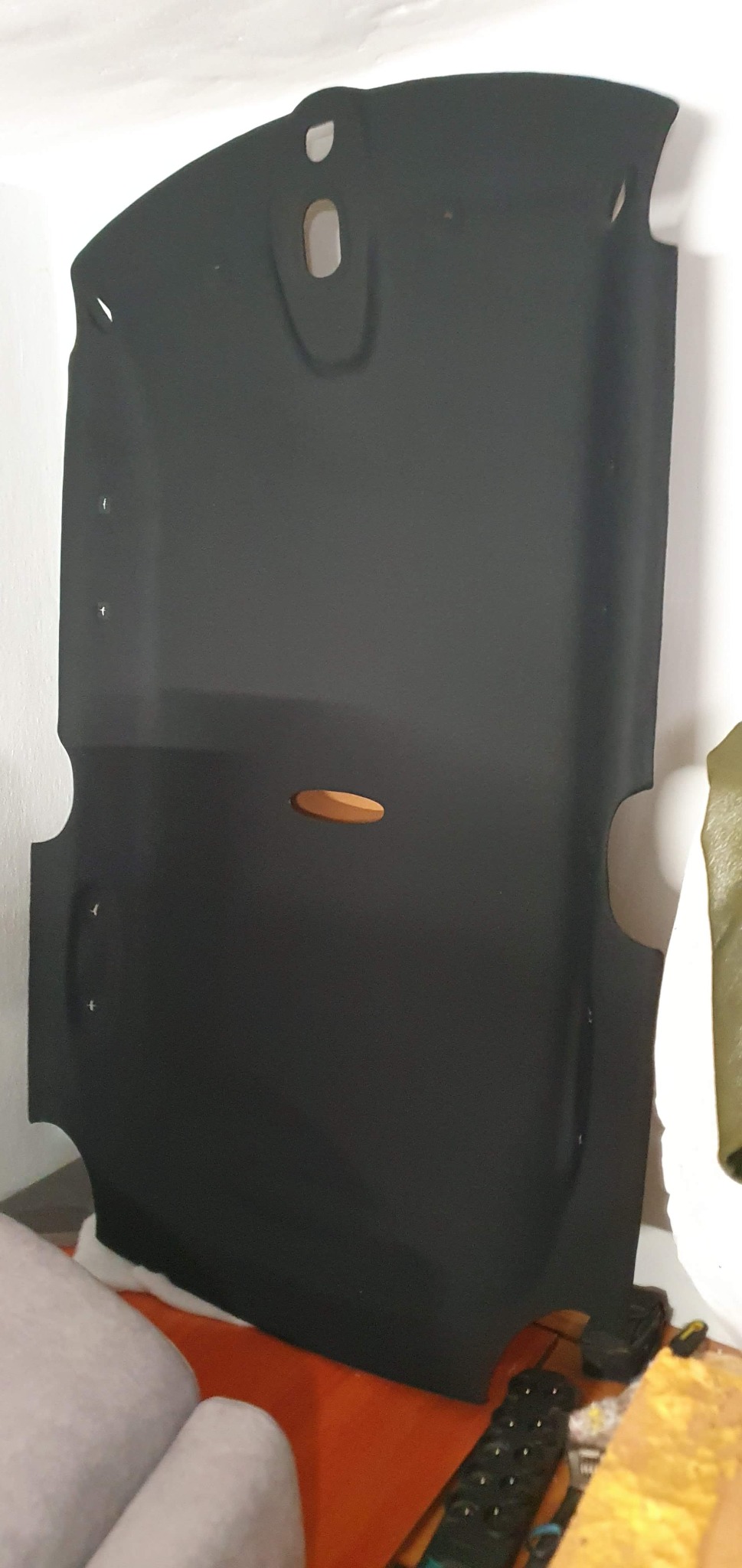 Czarna podsufitka samochodowa z wyciętymi otworami, oparta o białą ścianę, ukazująca detale tapicerki i kształt elementu przed montażem.