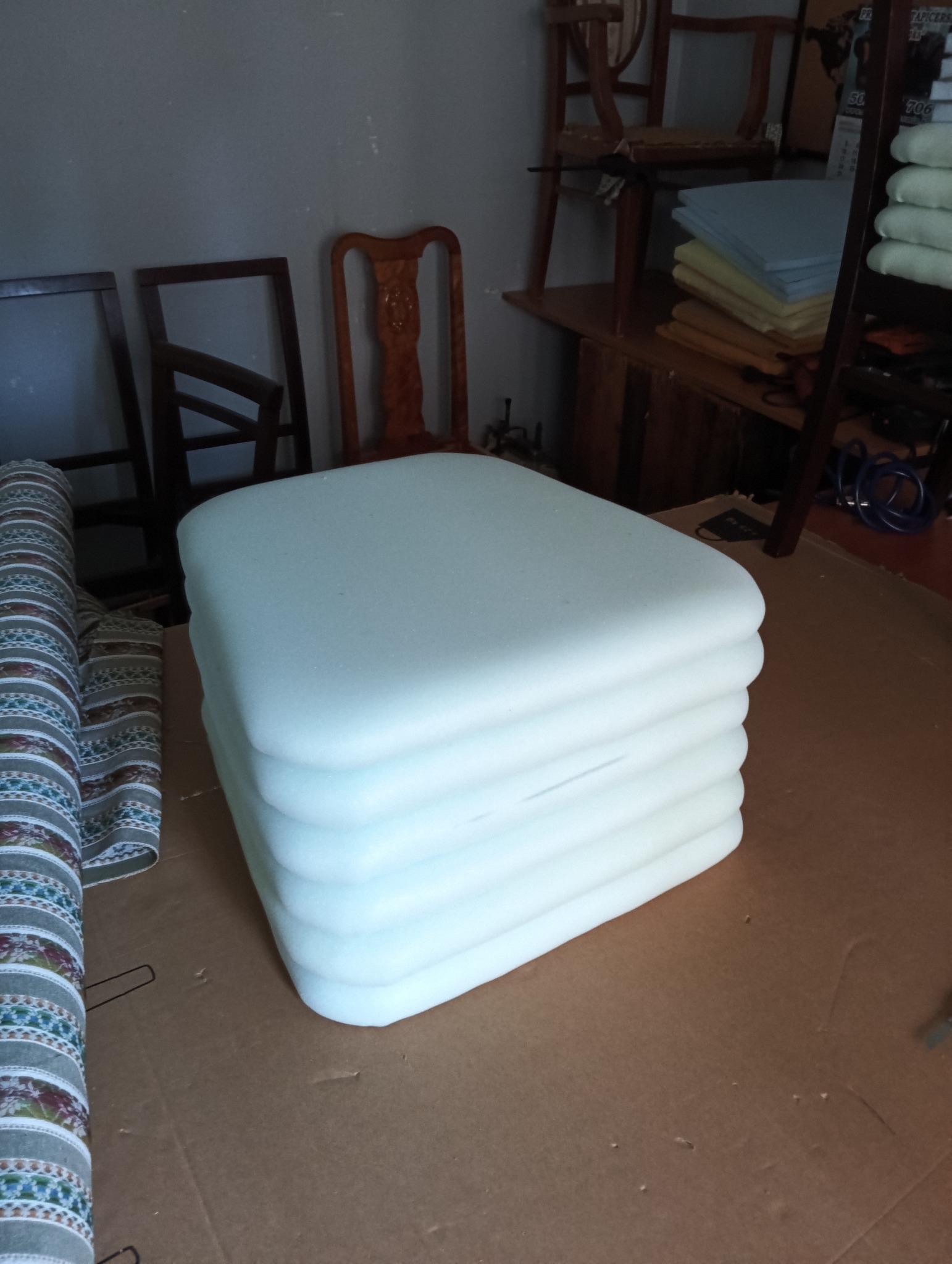 Stos piankowych siedzisk o zaokrąglonych krawędziach, obok zwinięta tkanina obiciowa w tle widoczne ramy krzeseł. Przygotowanie do prac tapicerskich.