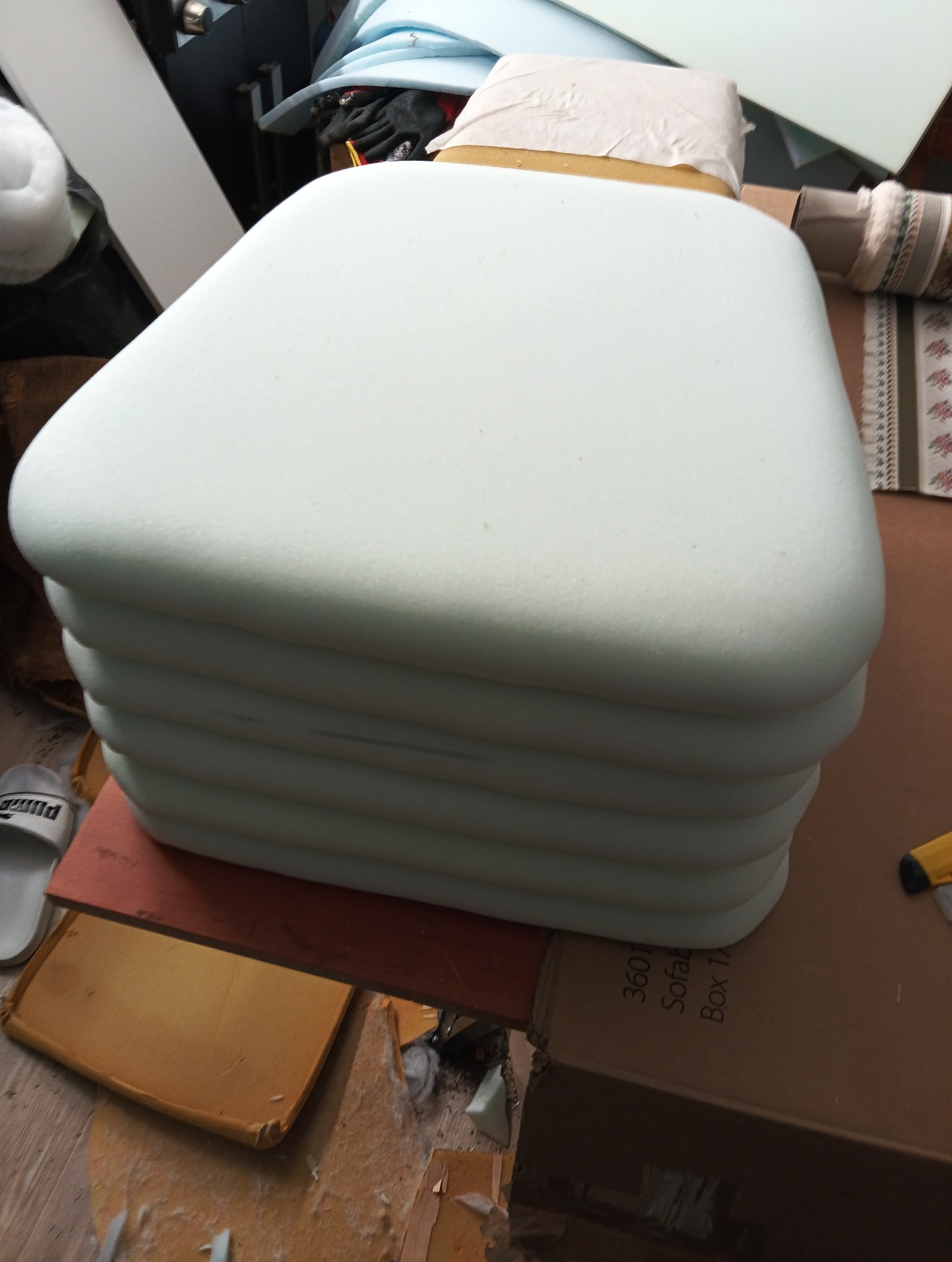 Stos jasnych, piankowych siedzisk o zaokrąglonych rogach, prawdopodobnie do mebli tapicerowanych, widoczny w warsztacie tapicerskim.