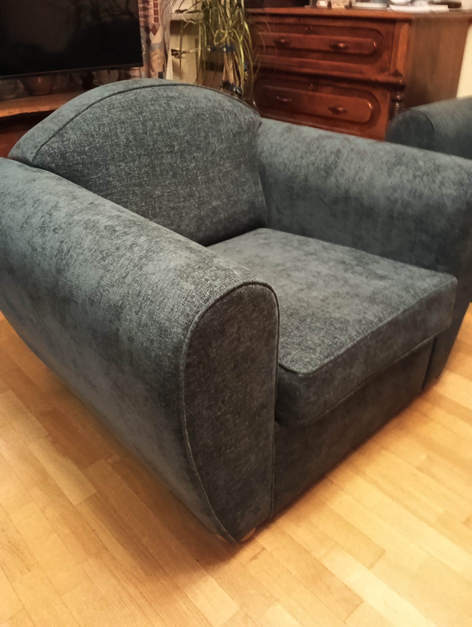 Szary fotel klubowy z zaokrąglonymi podłokietnikami na parkiecie, w tle komoda i roślina doniczkowa. Widoczne subtelne detale tapicerki i drewniane wykończenie.