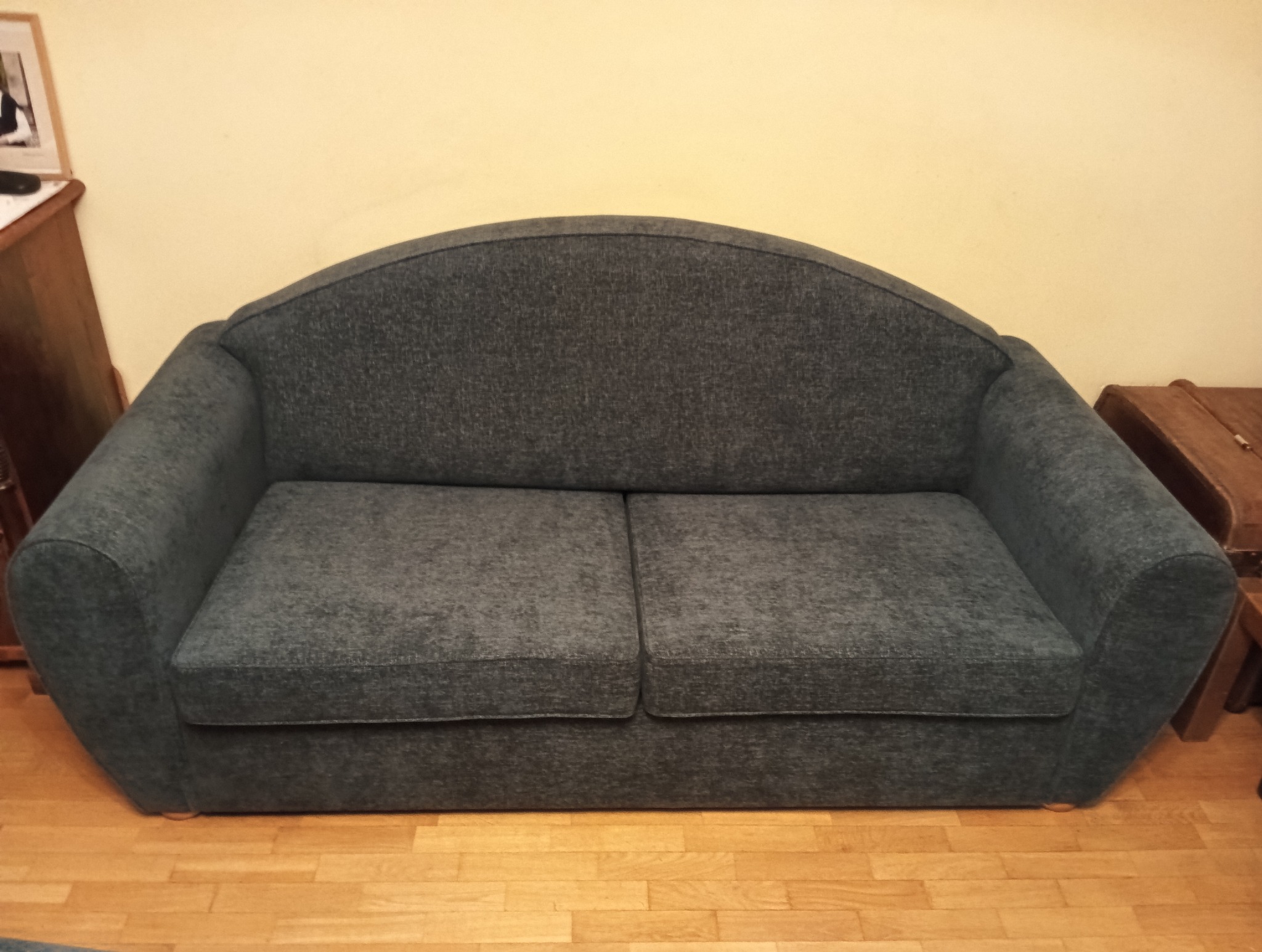 Dwuosobowa sofa w odcieniach szarości, z zaokrąglonym oparciem i podłokietnikami, stoi na drewnianej podłodze. Obok widoczny fragment drewnianej komody.