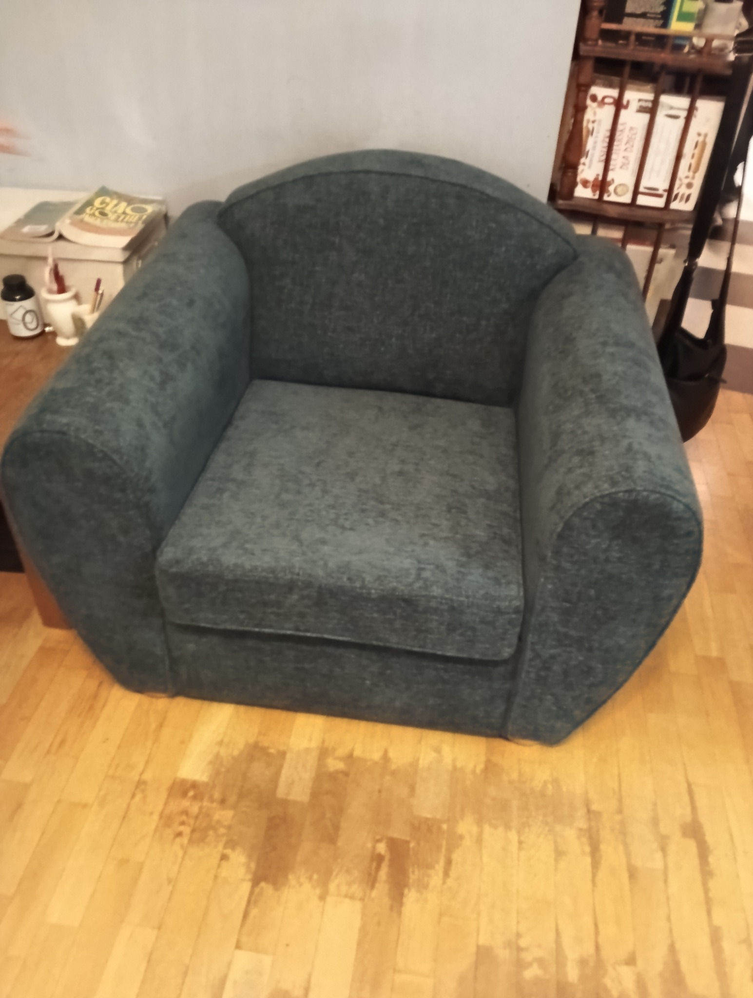 Szary fotel z zaokrąglonym oparciem na drewnianej podłodze. Widoczne książki i półka w tle. Ujęcie z bliska, prezentujące detale tapicerki i formę mebla.