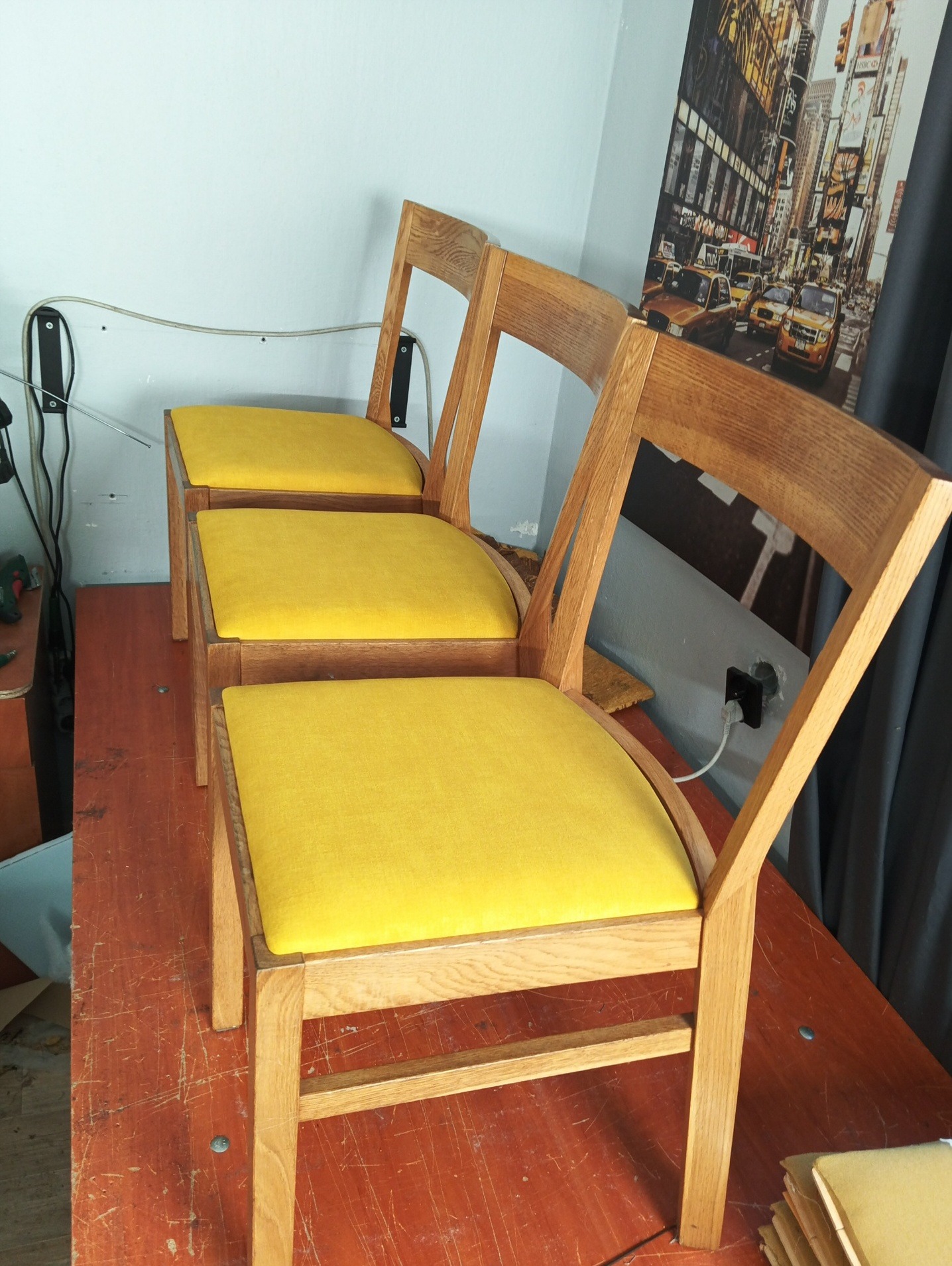 Trzy drewniane krzesła z żółtymi siedziskami ustawione w rzędzie, widoczne na tle ściany i obrazu z motywem miejskim. Krzesła po renowacji.