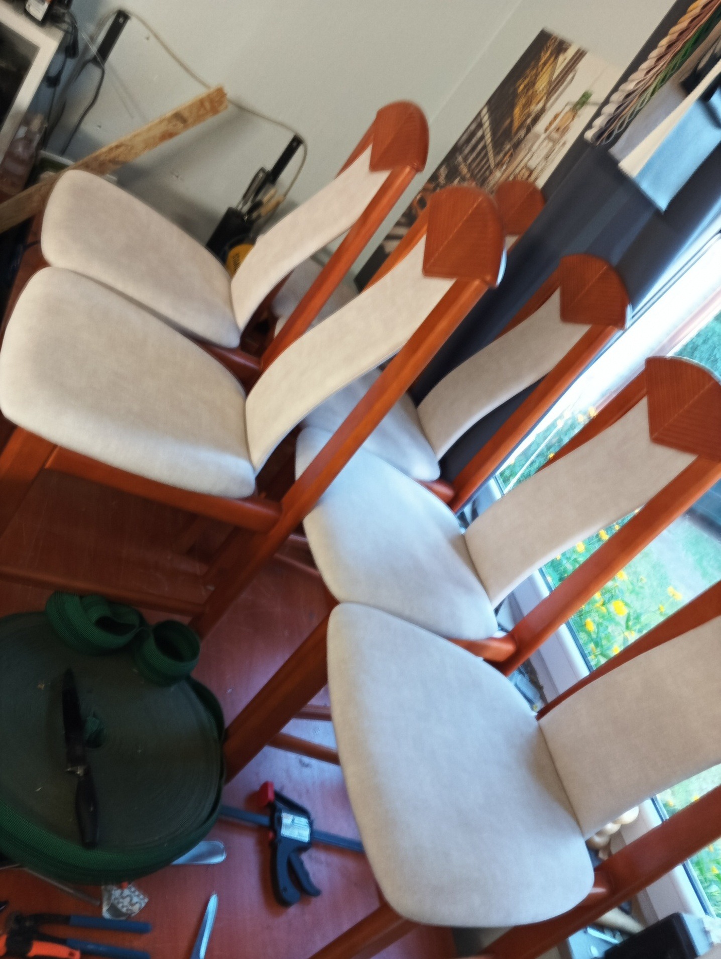 Odnowione krzesła z drewnianą ramą i jasną tapicerką, ustawione w rzędzie w pracowni. Widoczne narzędzia tapicerskie. Usługi tapicerskie w Warszawie.