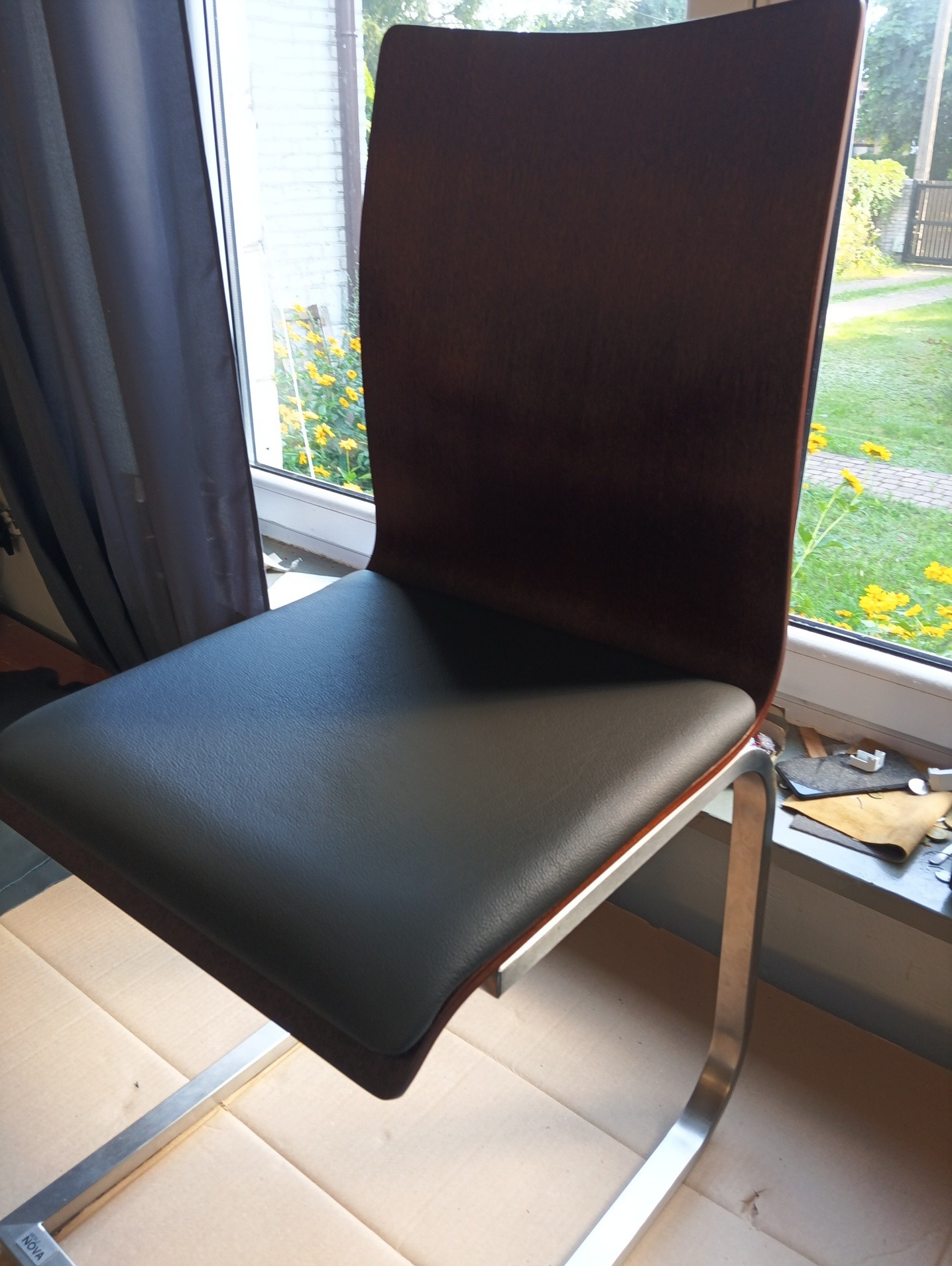 Krzesło z ciemnobrązowym oparciem i czarnym siedziskiem, na metalowych nogach. W tle okno z widokiem na ogród i narzędzia tapicerskie.