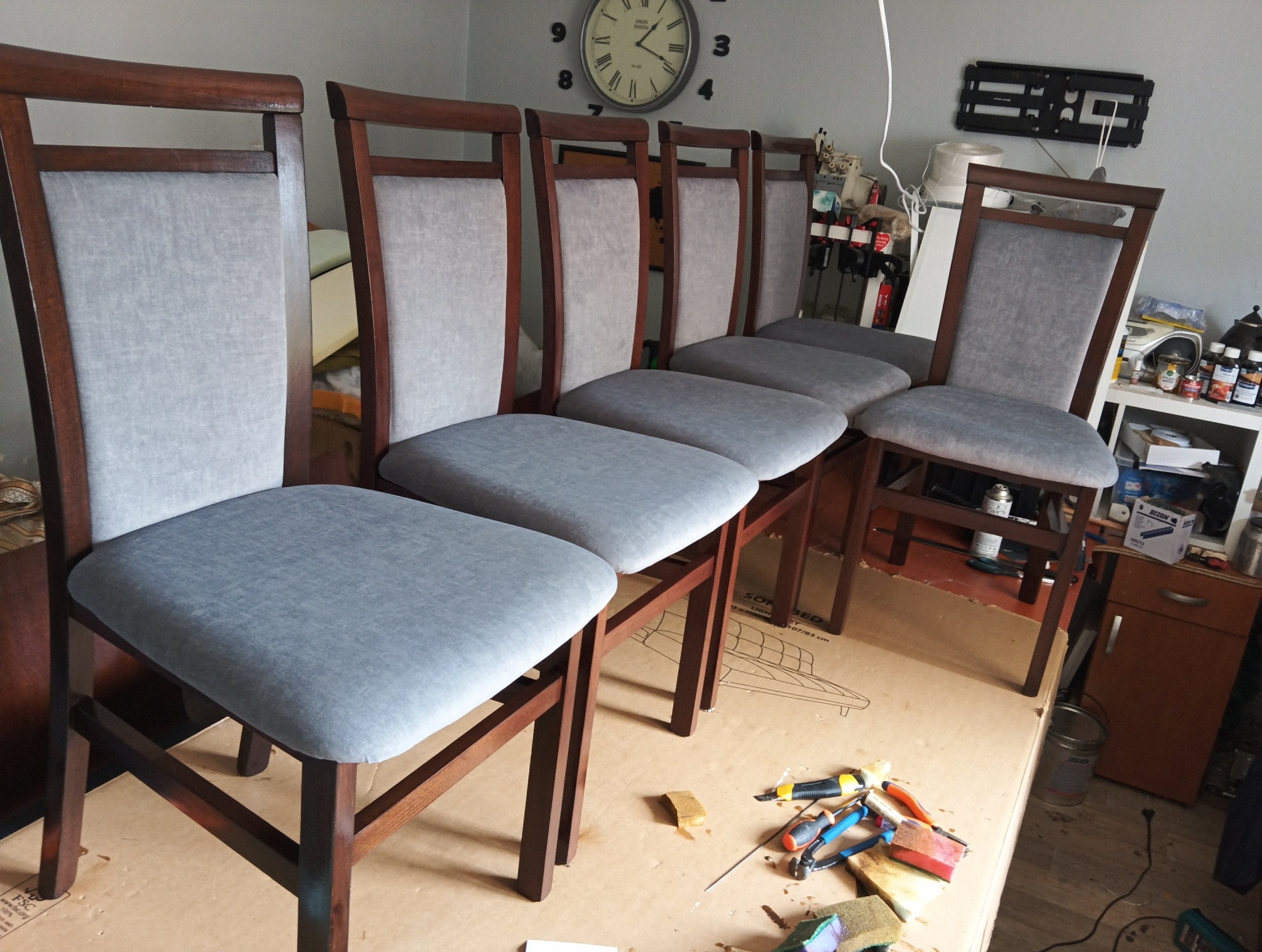 Rząd sześciu krzeseł tapicerowanych szarą tkaniną, z drewnianymi ramami w odcieniach brązu. Na pierwszym planie narzędzia tapicerskie.