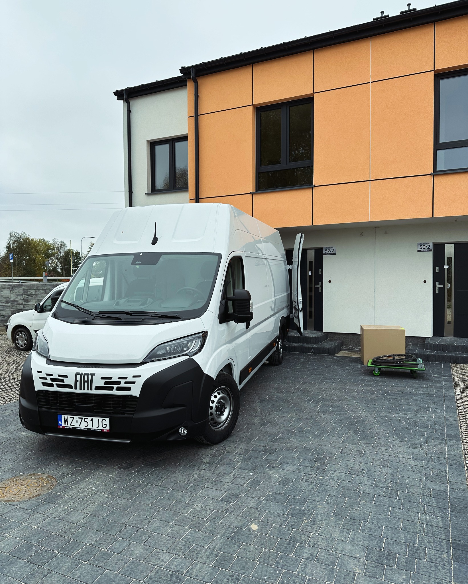 Biały samochód dostawczy Fiat z otwartymi drzwiami zaparkowany przed nowoczesnym budynkiem z pomarańczową elewacją, obok karton na wózku transportowym.