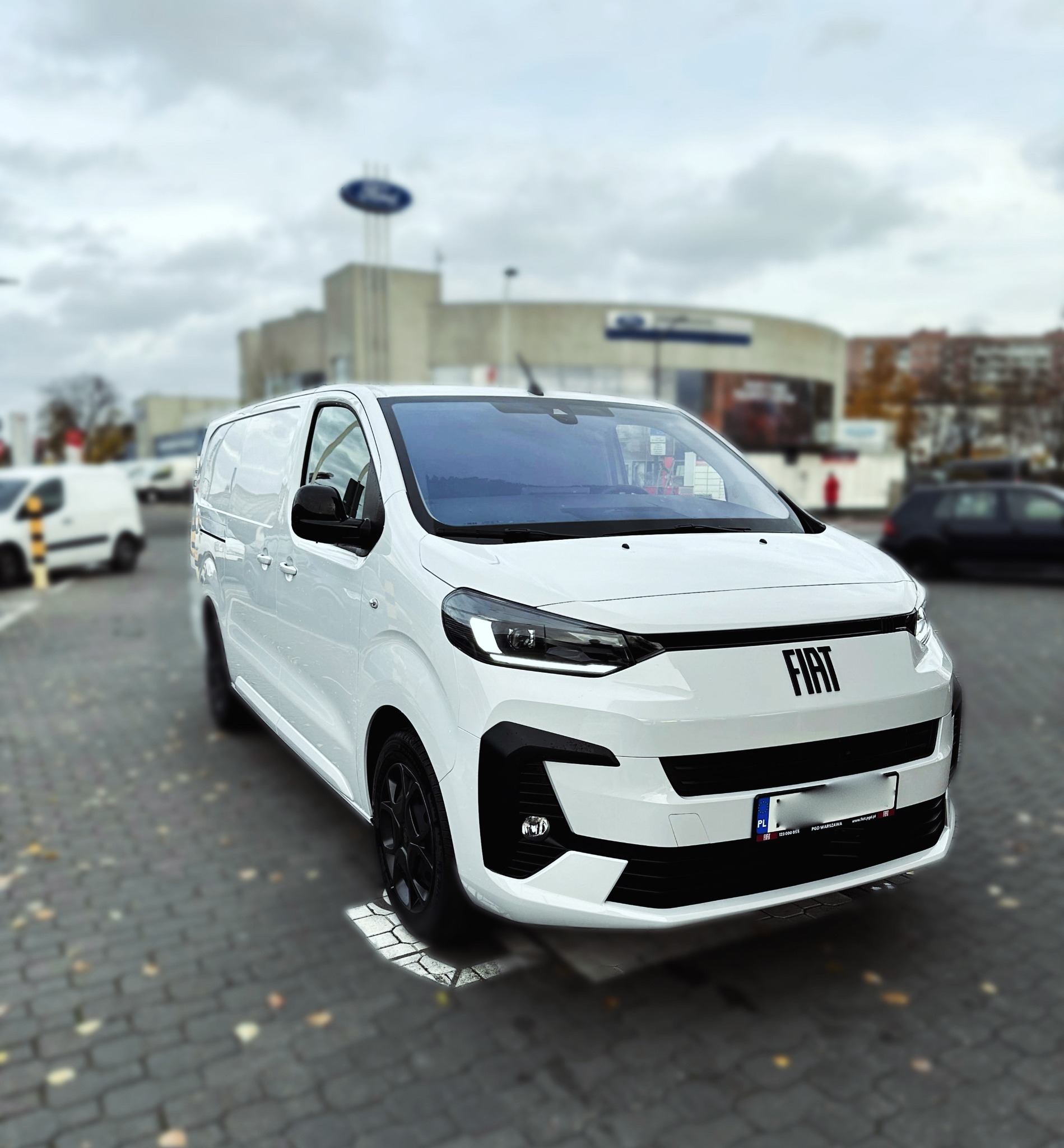 Biały samochód dostawczy marki Fiat z czarnymi felgami na parkingu z kostki brukowej, na tle budynku z logo Forda.