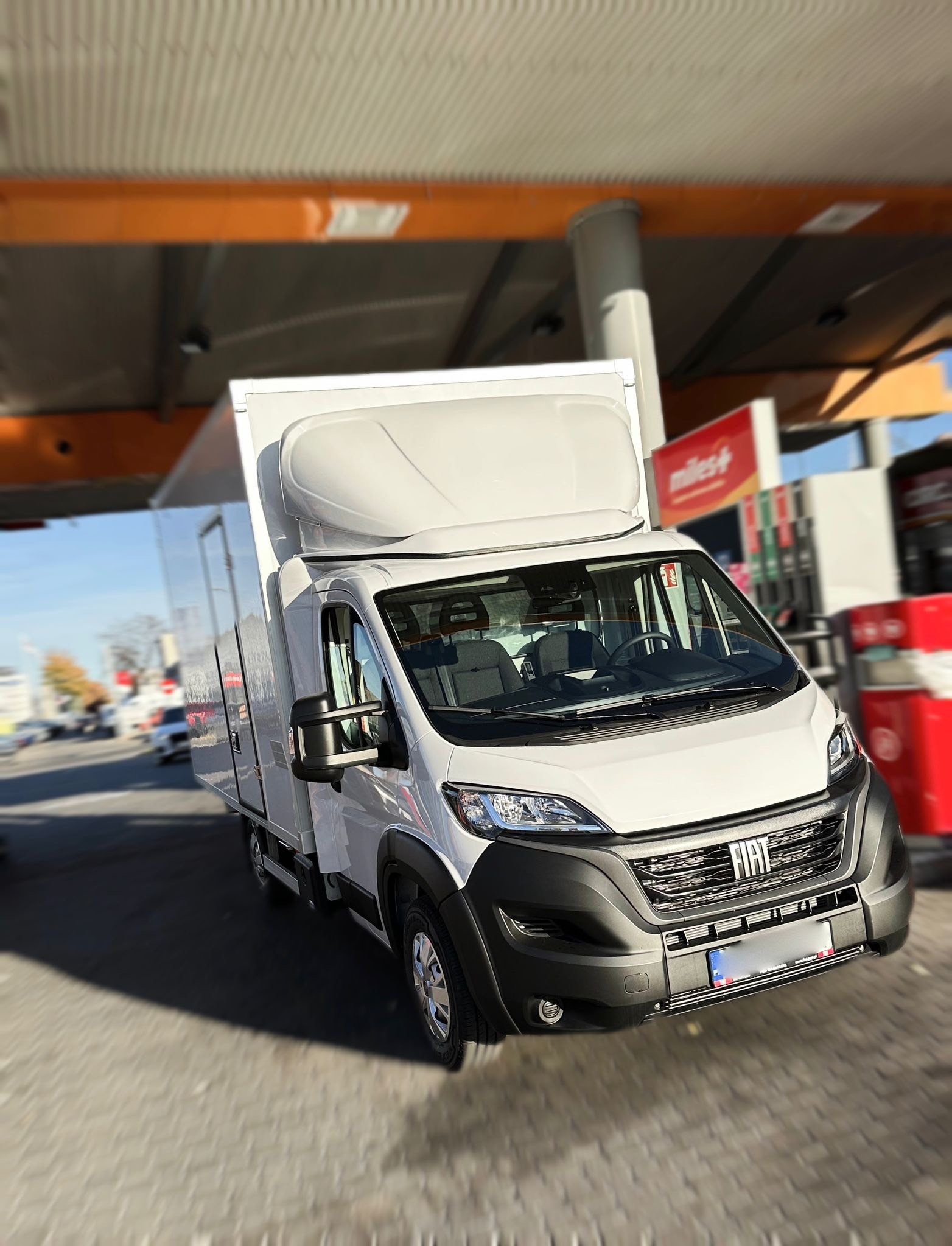 Biały samochód dostawczy Fiat Ducato z wysoką zabudową, zaparkowany na stacji benzynowej w słoneczny dzień.