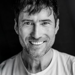 COSMO MEDIA ŁUKASZ KLIMEK - Czarno-biały portret uśmiechniętego mężczyzny z krótką brodą i ciemnymi włosami na ciemnym tle, z widocznymi zmarszczkami wokół oczu.