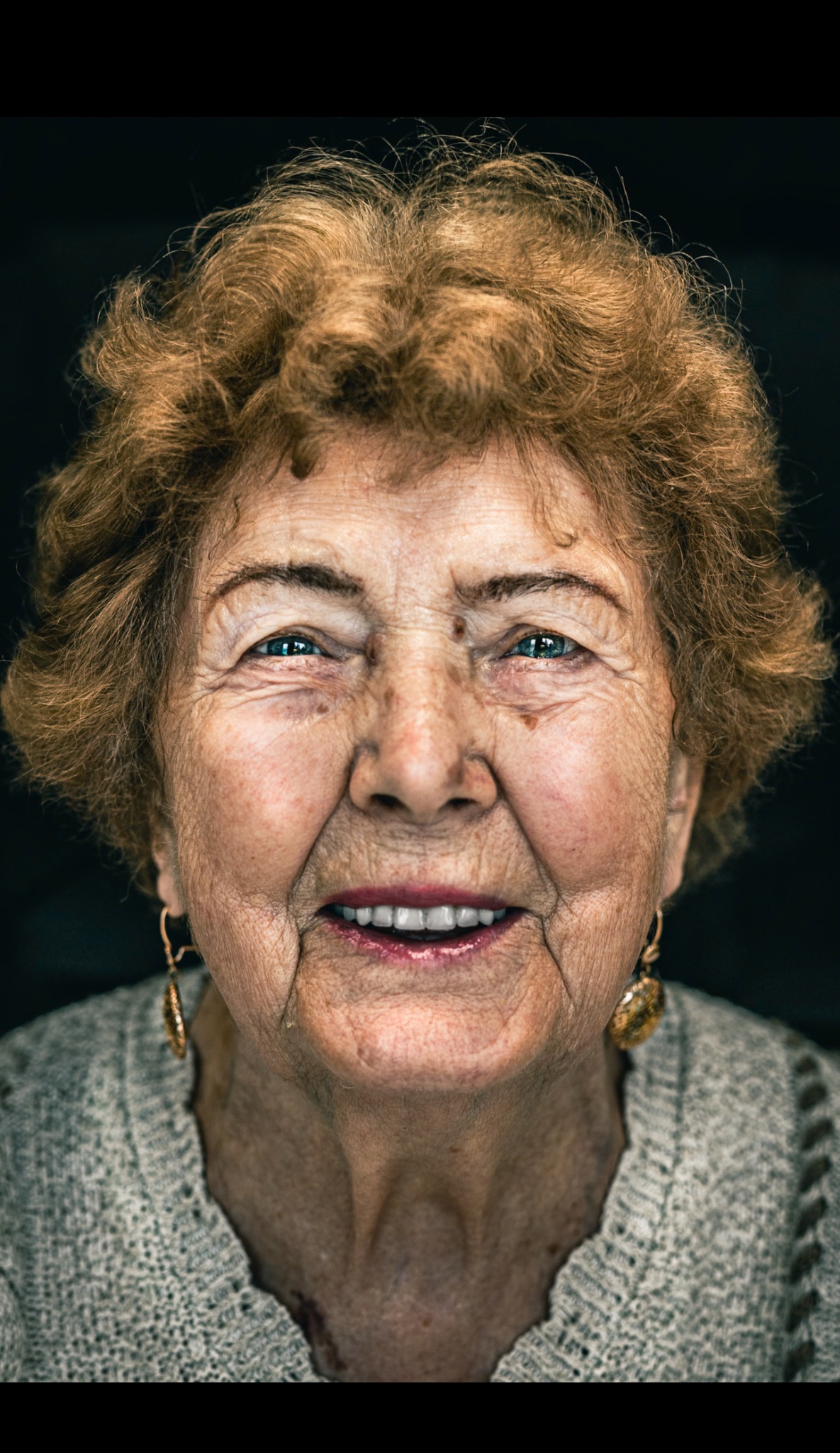 Portret uśmiechniętej starszej kobiety z kręconymi włosami w ciepłym odcieniu blondu, z widocznymi zmarszczkami i złotymi kolczykami, na ciemnym tle.