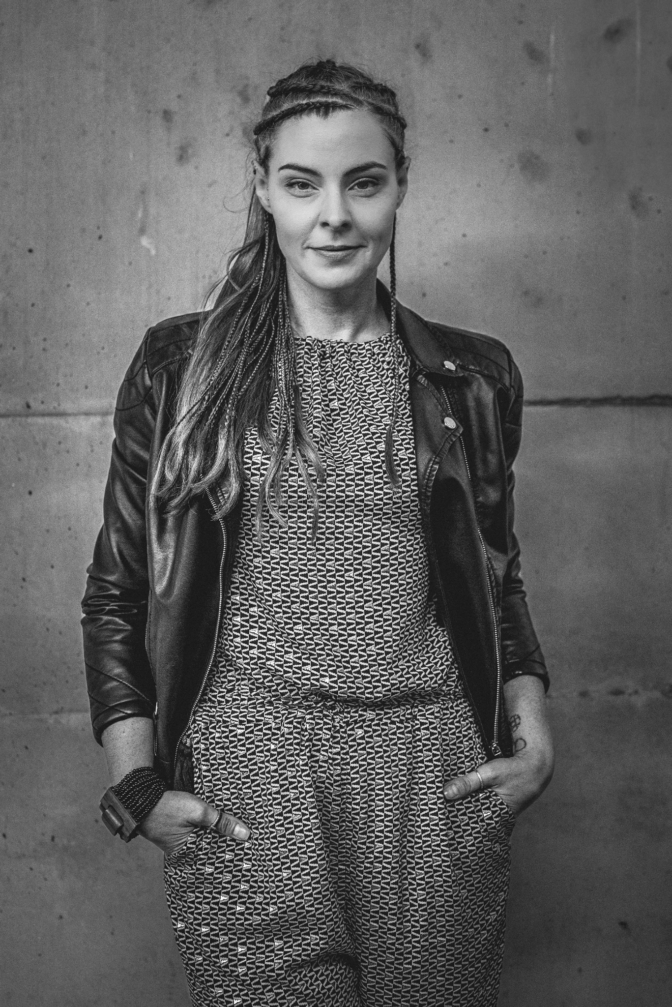 Czarno-biały portret kobiety w skórzanej kurtce, z zaplecionymi włosami i subtelnym uśmiechem na tle surowej, betonowej ściany. Uwagę przykuwa wzorzysty kombinezon.