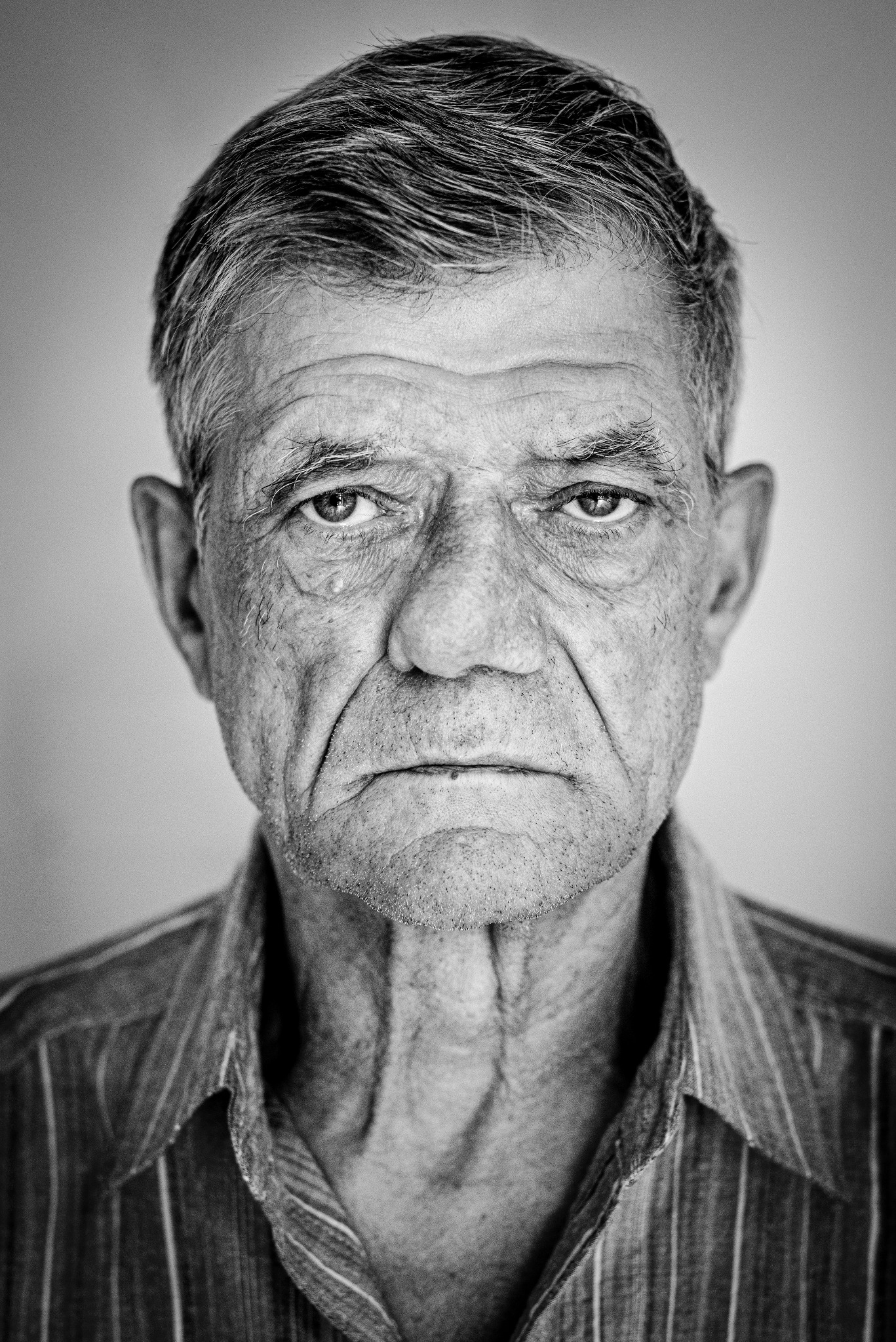 Czarno-biały portret starszego mężczyzny z wyraźnymi zmarszczkami, patrzącego wprost w obiektyw, z bliska, w koszuli w paski i neutralnym tle.