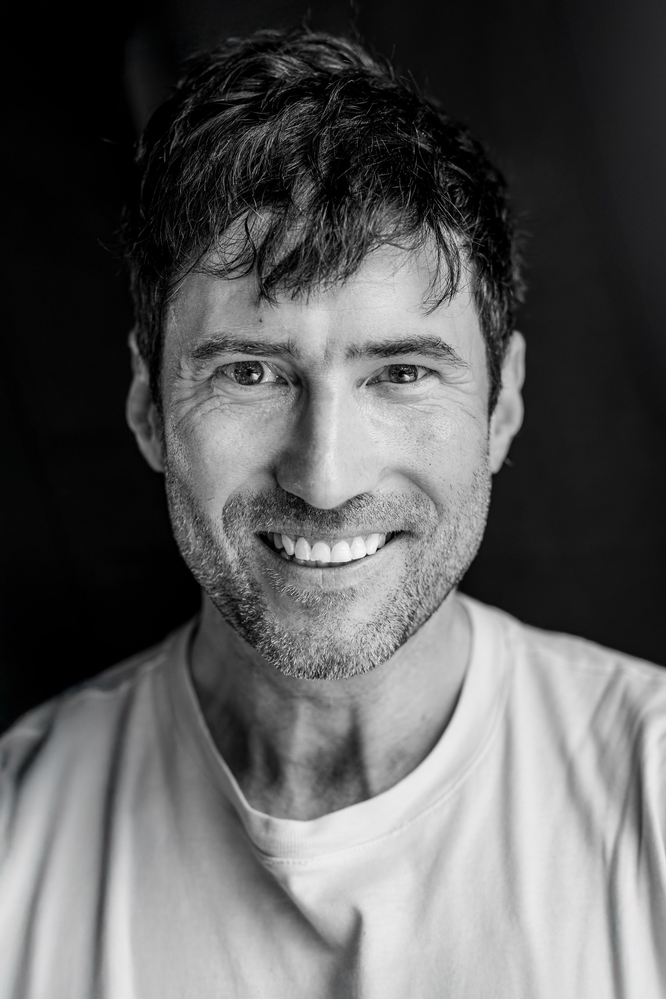 Czarno-biały portret uśmiechniętego mężczyzny z krótką brodą i ciemnymi włosami na ciemnym tle, z widocznymi zmarszczkami wokół oczu.