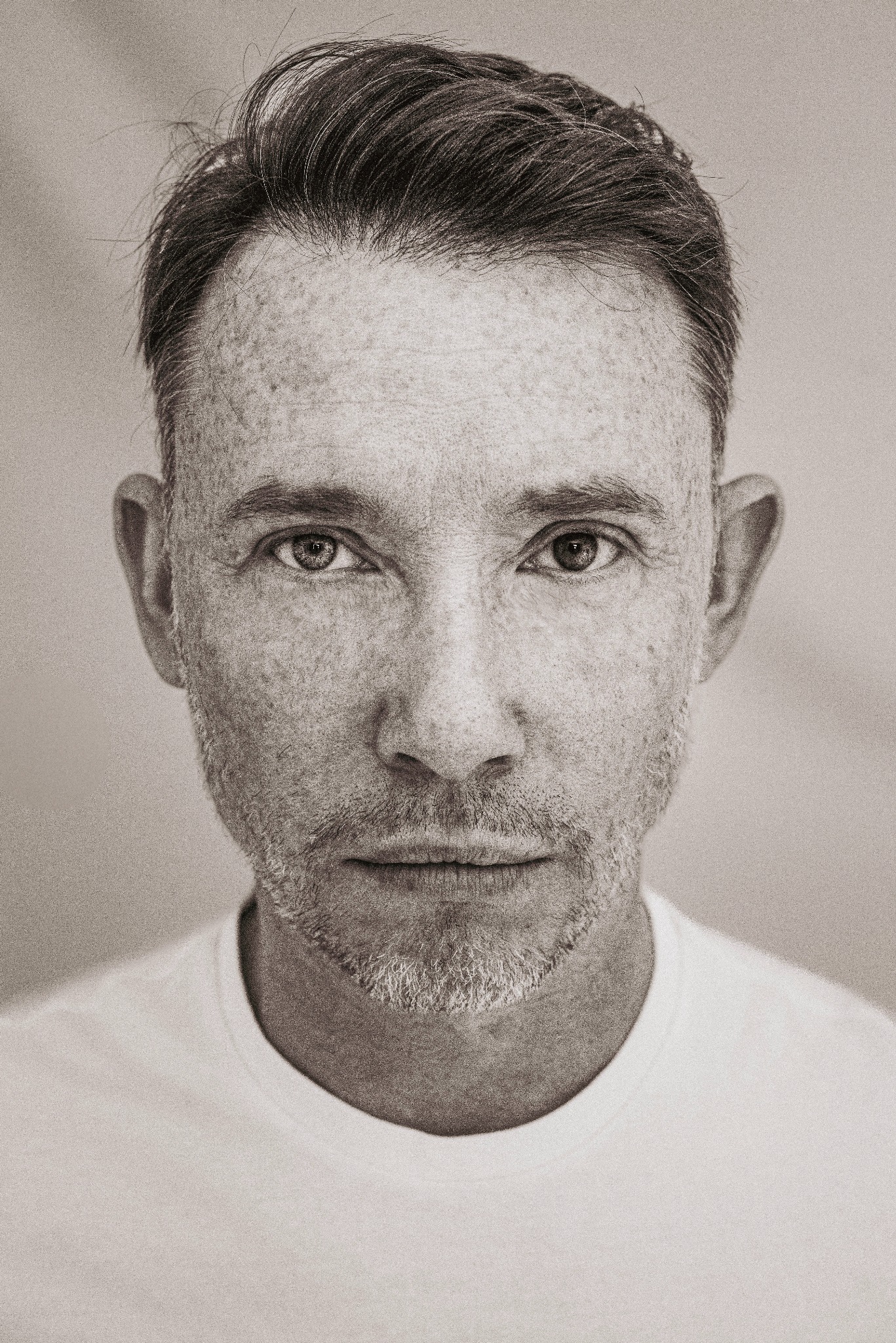 Czarno-biały portret mężczyzny z brodą i piegami, na wprost, w studiu w Warszawie. Neutralny wyraz twarzy, biała koszulka, jednolite tło.