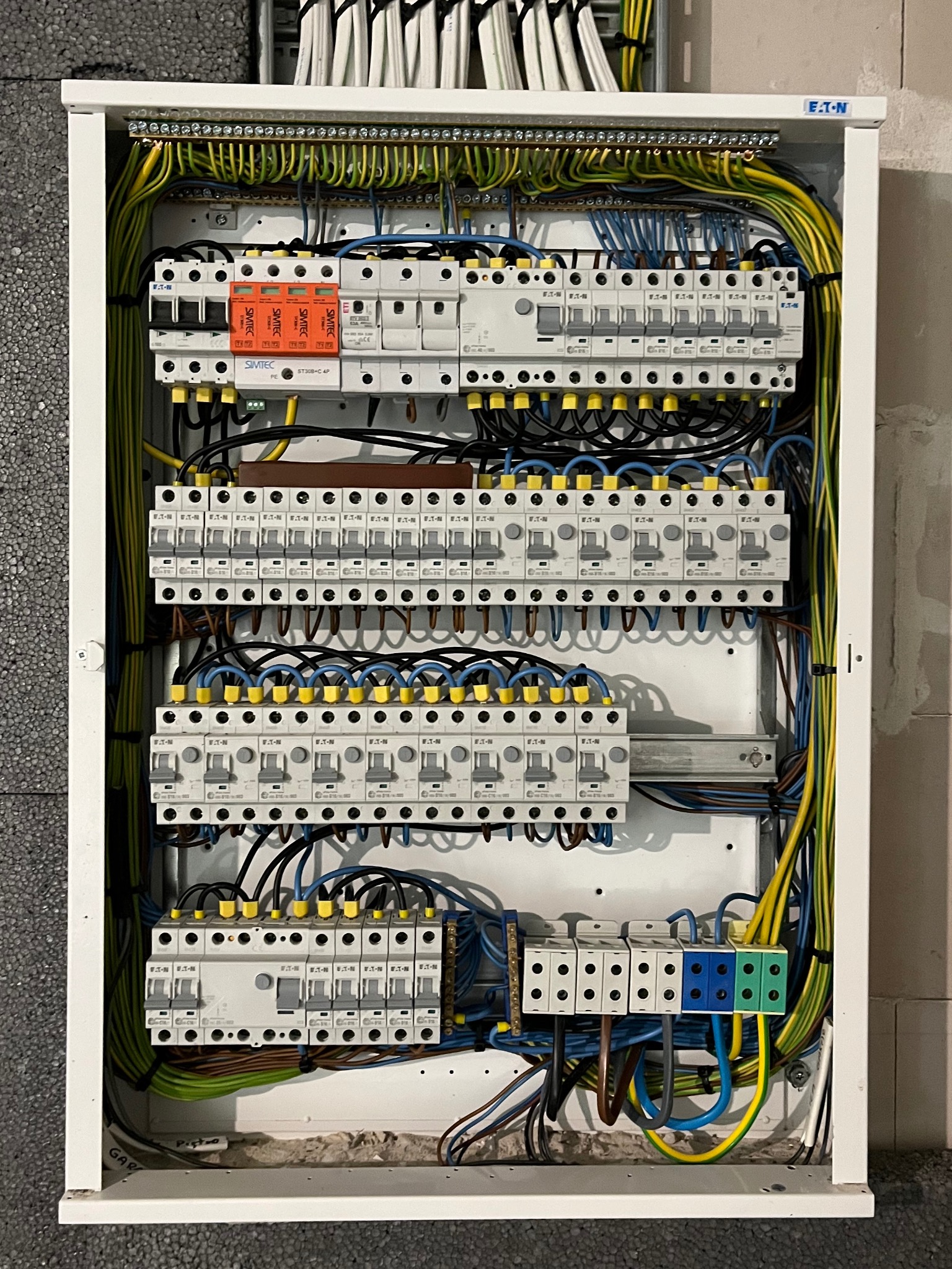 Otwarta skrzynka elektryczna z widocznymi bezpiecznikami, przewodami w różnych kolorach (żółto-zielone, niebieskie, czarne, brązowe) oraz listwami zaciskowymi; widoczne elementy marki Eaton i Simtec.