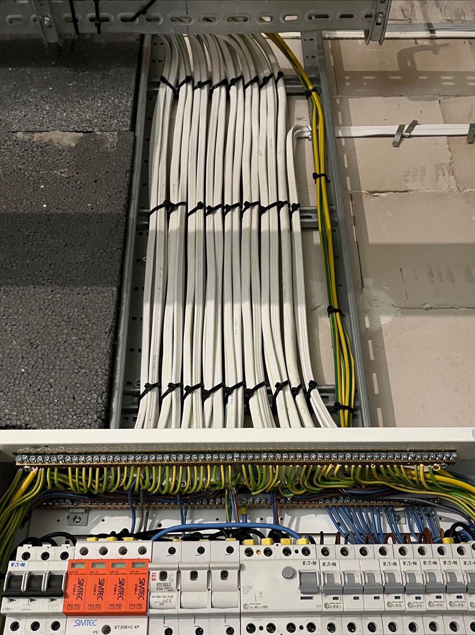 Równolegle ułożone białe przewody elektryczne, zabezpieczone czarnymi opaskami zaciskowymi, prowadzone w metalowej korycie kablowej. Widoczna rozdzielnica elektryczna z bezpiecznikami...