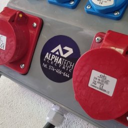 AlphaTech Electric - Szara skrzynka rozdzielcza z trzema niebieskimi gniazdami 16A i dwoma czerwonymi gniazdami 32A, zamontowana na białej ścianie. Widoczny podłączony czarny kabel i naklejka firmy 'ALPHATECH ELECTRIC'.