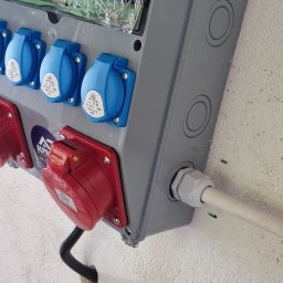 AlphaTech Electric - Szara skrzynka rozdzielcza z trzema niebieskimi i jednym czerwonym gniazdem przemysłowym, zamontowana na jasnej ścianie, z zabezpieczeniami pod pogniecioną folią ochronną.
