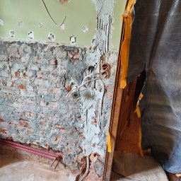 Usługi Elektryczne Arkadiusz Kowalewski - Ściana w trakcie remontu z odsłoniętą instalacją elektryczną i fragmentem muru ceglanego, zabezpieczenie folią malarską przy framudze drzwi.